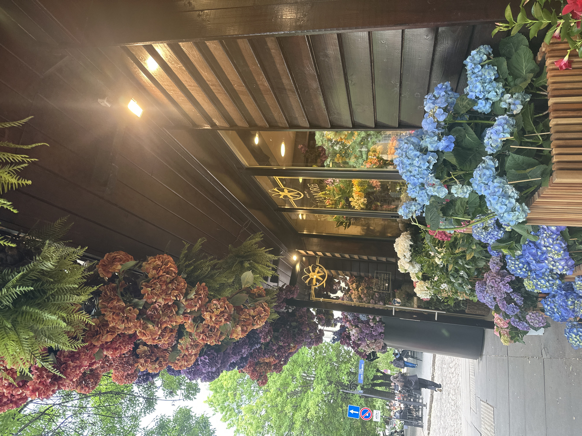 The image shows the exterior of a flower shop in Sofia, Bulgaria. The shop has a dark wood awning and large glass windows. A sign above the window reads "L'AMORE STYLE B". The storefront is decorated with an abundance of colorful hydrangeas and greenery. On the street, pedestrians walk past, and a blue street sign is visible.