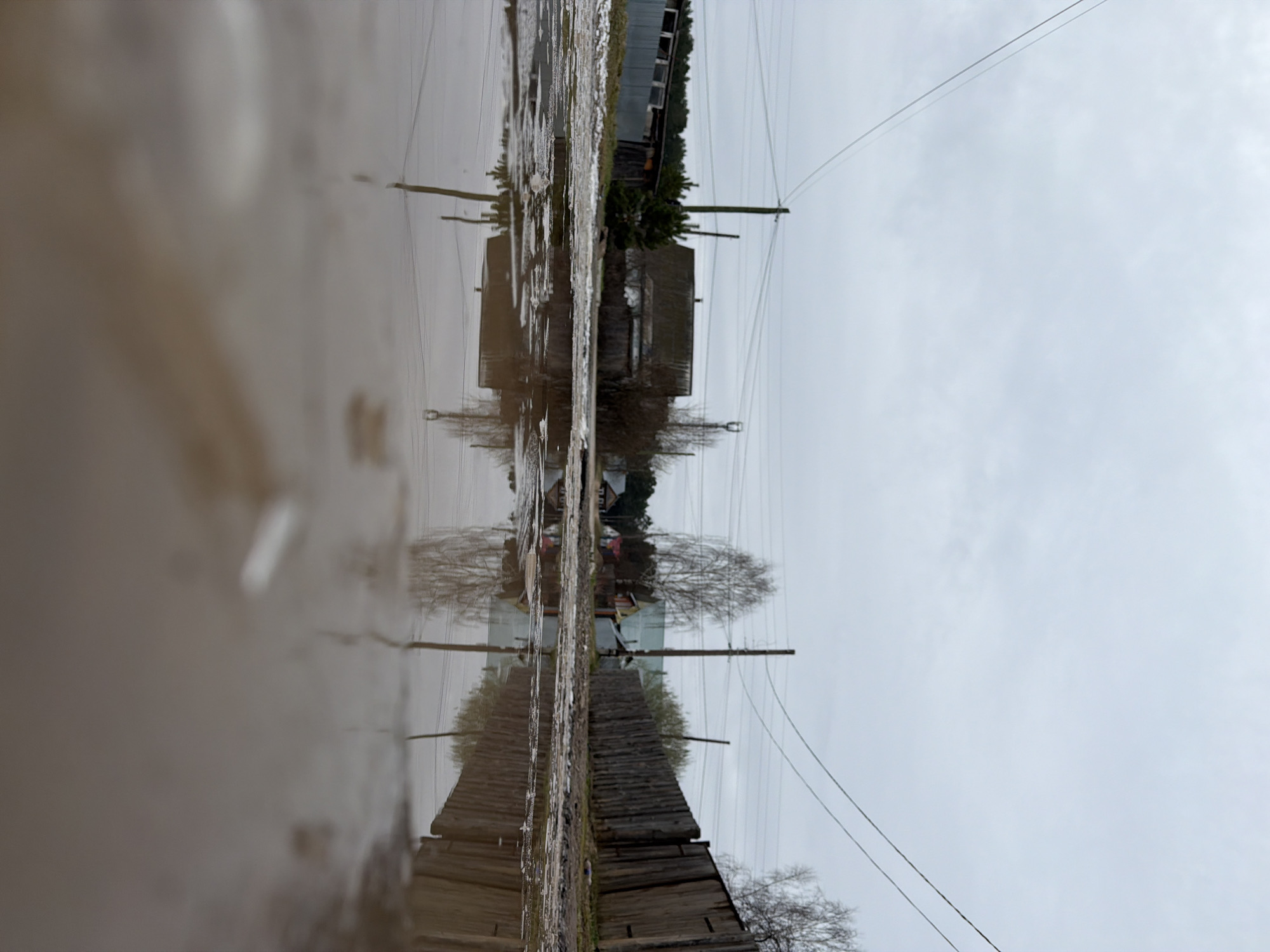 The image is a reflection in a puddle. The puddle reflects a scene in Chaikovsky, Russia, featuring a weathered wooden structure with a sloping roof, adjacent to a fence made of uneven wooden planks. The image also shows utility poles with wires running across the top of the frame. In the background, there's a glimpse of a building with multiple windows and trees. The sky appears overcast.