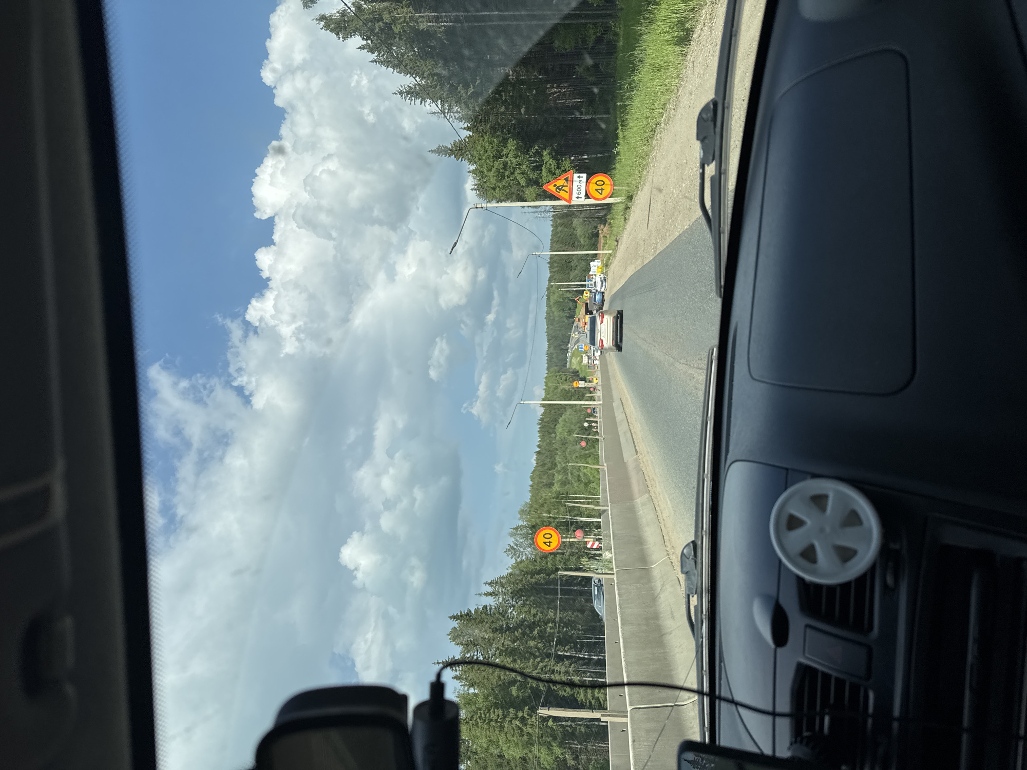 The image captures a view from the driver's seat of a car traveling on a highway with a concrete barrier on the left and a dense forest on the right. The sky is blue with scattered white clouds. Road signs are visible, including a speed limit sign indicating "40" and another sign with a construction worker symbol and "600m." Several cars are on the road ahead. The dashboard of the car is visible in the foreground, with a small, white, flower-shaped object attached to a vent. The road appears to be under construction or repair, with barriers and possibly machinery visible in the distance. The scene suggests a typical highway environment in a rural or semi-rural area, likely in Russia given the context of Nytva.