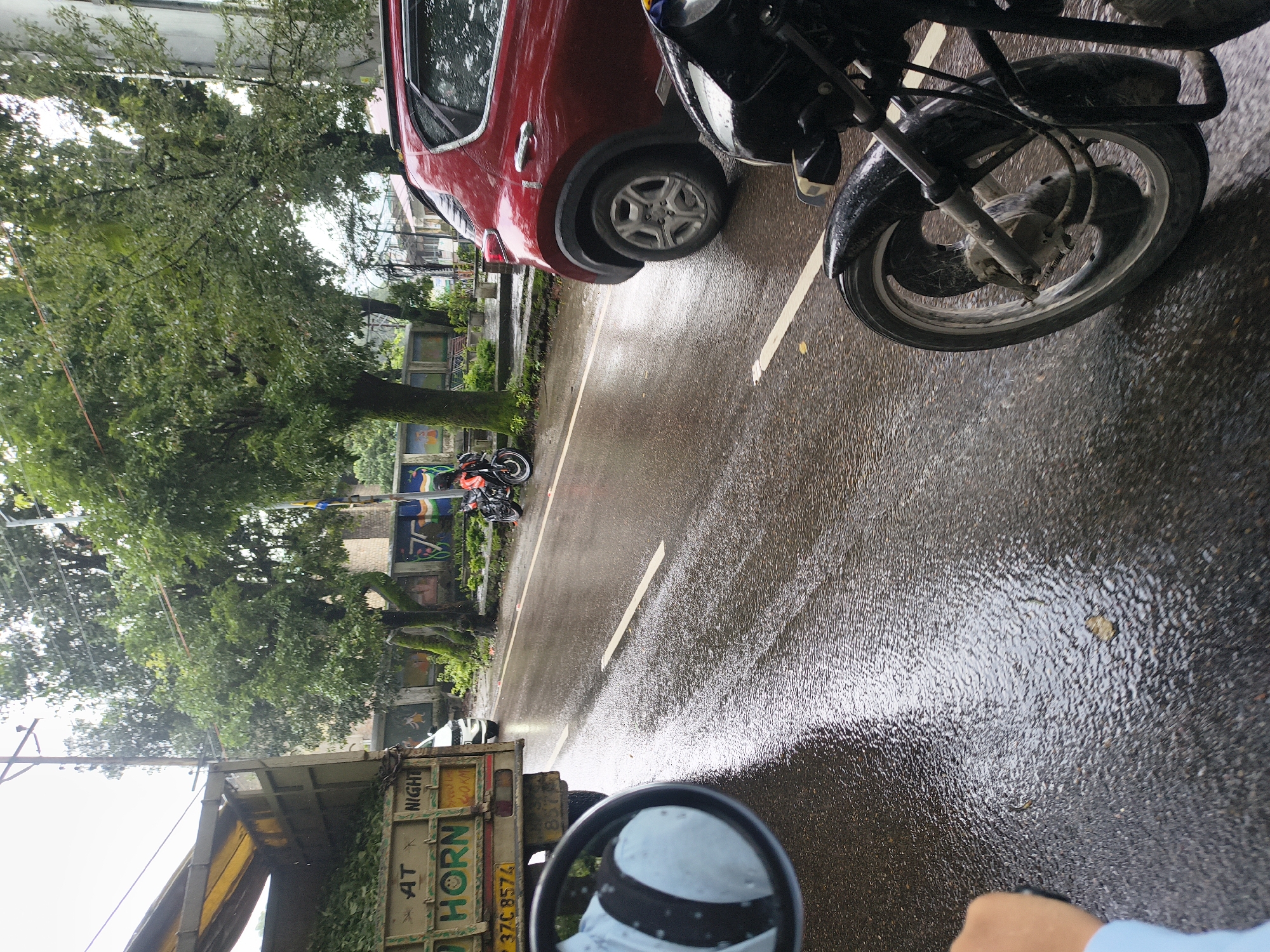 The image depicts a street scene in Dharamshala, India, during what appears to be monsoon season, with the road wet and reflecting the overcast sky. In the foreground, the front wheel and handlebars of a black motorcycle are visible, indicating the perspective of the photographer. To the left, a side-view mirror reflects a person's arm and shoulder, wearing a light blue shirt with a black stripe. Further back, a light-colored truck is parked on the side of the road, loaded with green foliage. The truck has "HORN" written on its side. A white car is also present behind the truck. A red SUV is parked on the right side of the road. In the background, several trees line the street, and a wall with colorful murals is visible. Two motorcycles are parked near the wall. The presence of rain or recent rain is evident from the wet road surface and water droplets on some surfaces.