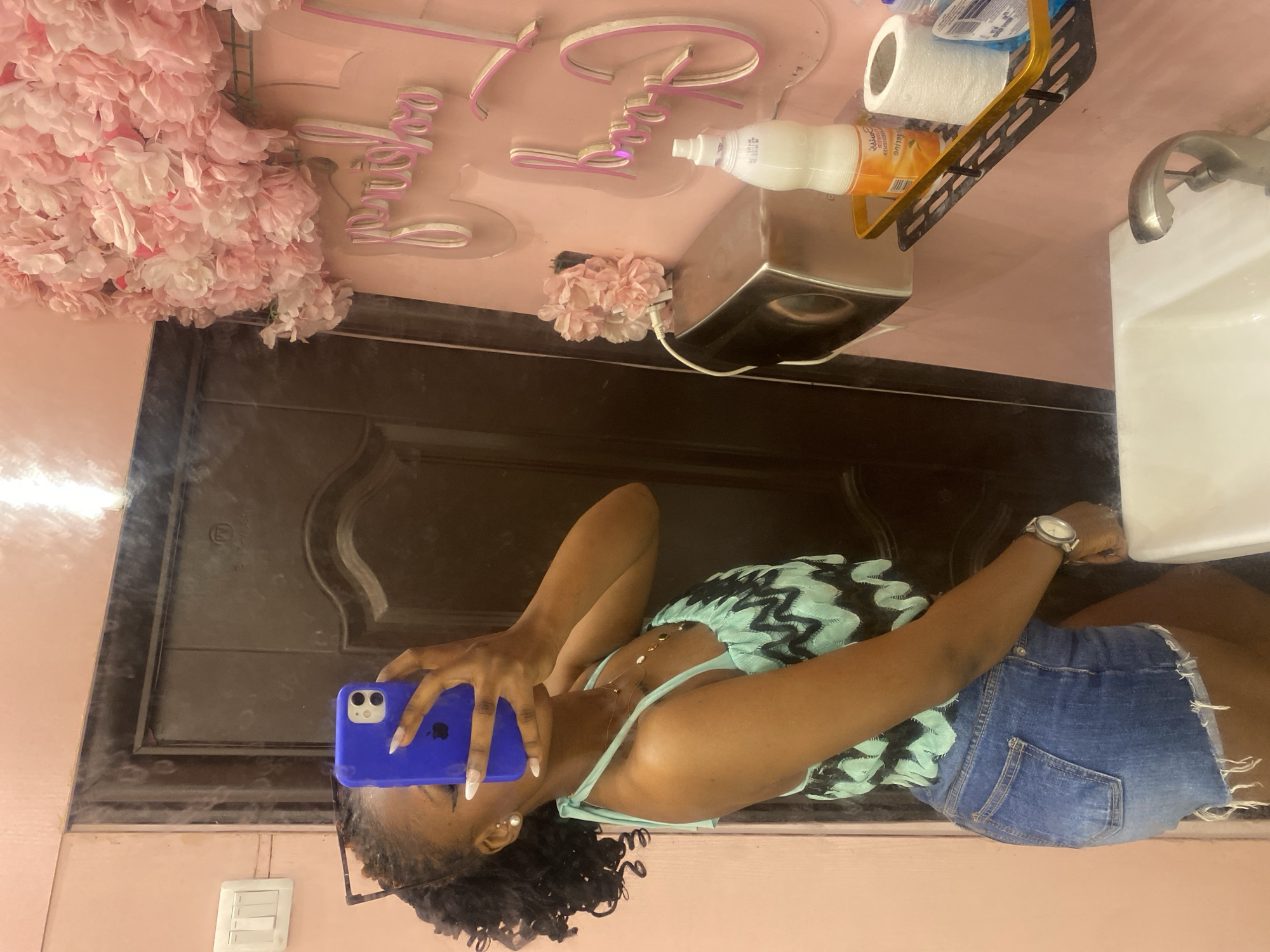 A young woman with braided hair, wearing a blue phone case over her face, a light blue and black zig-zag patterned halter top, and denim shorts, stands in a bathroom. She is holding her phone to take a selfie in the mirror. The bathroom has pink walls, a dark brown door, a white sink, a chrome faucet, and a shelf with toiletries including a bottle of lotion and a roll of toilet paper. There is a pink floral arrangement on the wall and a pink sign with illuminated text that reads "Bridal Shower". The location is specified as Howayi, Nigeria.