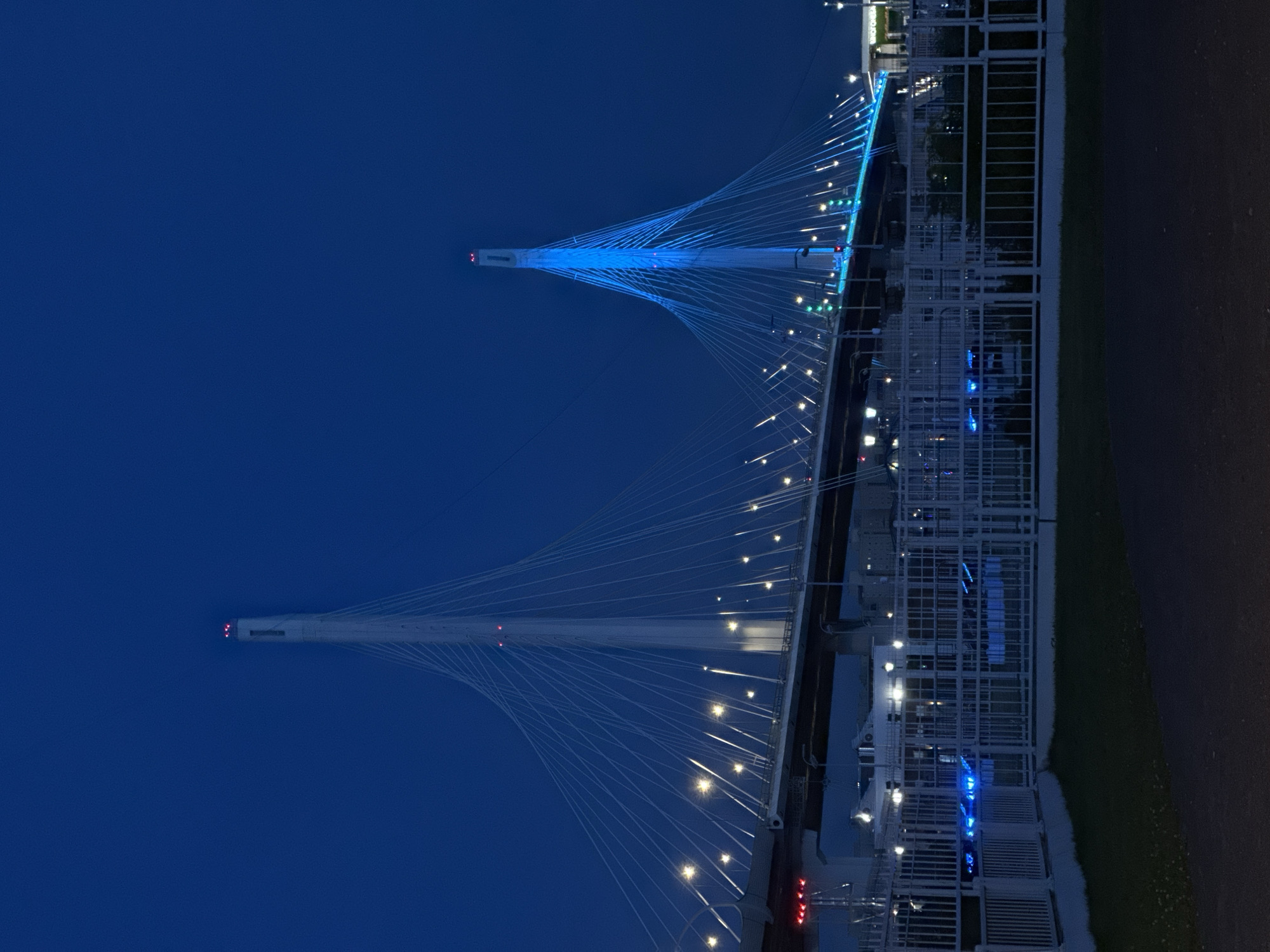 The image shows the Big Obukhovsky Bridge in St. Petersburg, Russia, at night. The bridge is a cable-stayed bridge illuminated with blue and white lights. Two tall pylons rise from the bridge, supporting a web of cables that hold up the roadway. The sky is a deep blue, and lights from the city are visible in the distance. A white metal fence runs along the foreground, separating the viewer from the bridge. The overall impression is one of a modern and majestic piece of infrastructure against a dark, twilight sky.