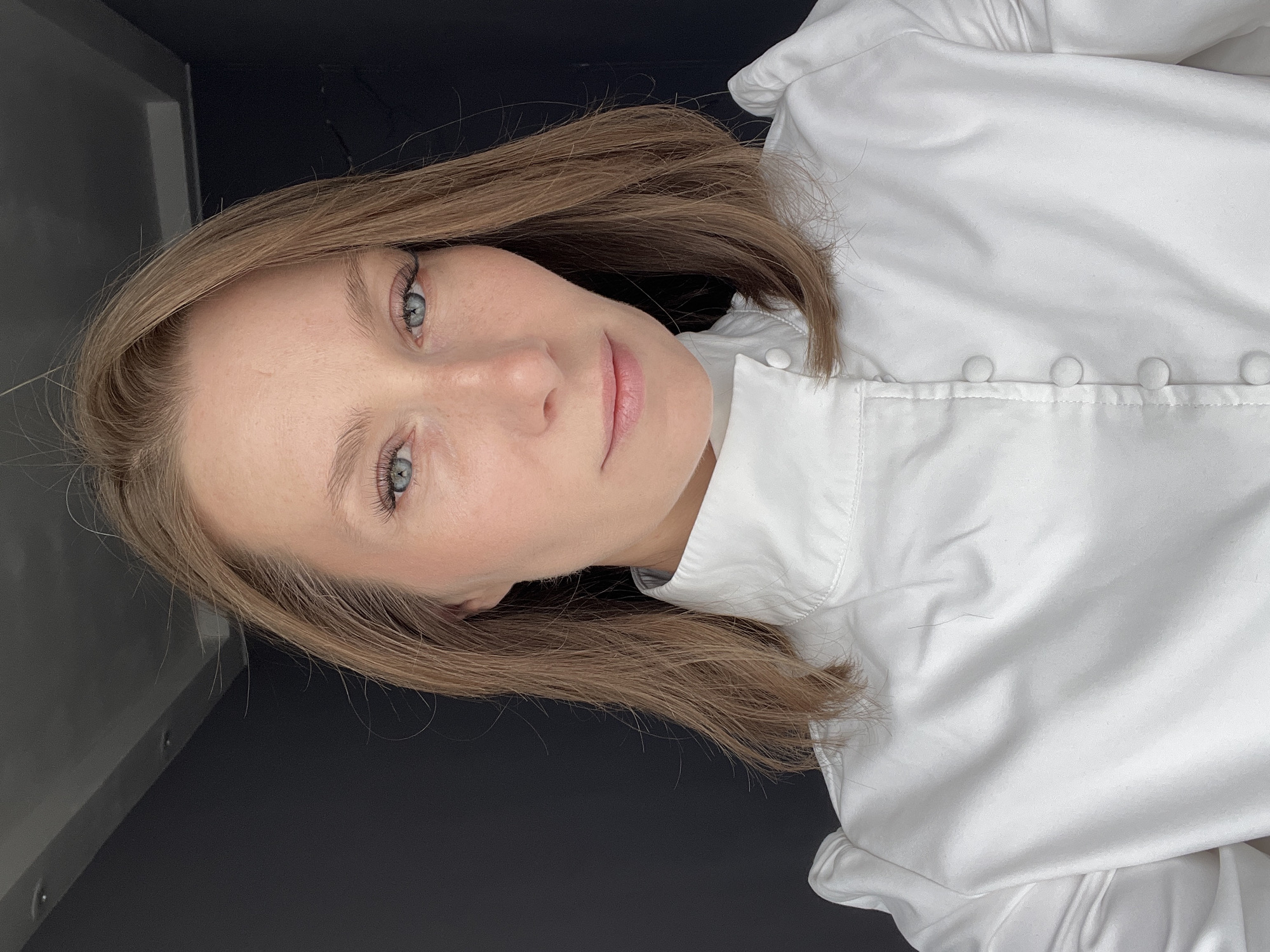 The image depicts a light-skinned woman with shoulder-length light brown hair and light-colored eyes wearing a white, collared button-down shirt. The background is dark, suggesting an interior setting. There is no information to determine that this image was taken in Taldom, Russia.