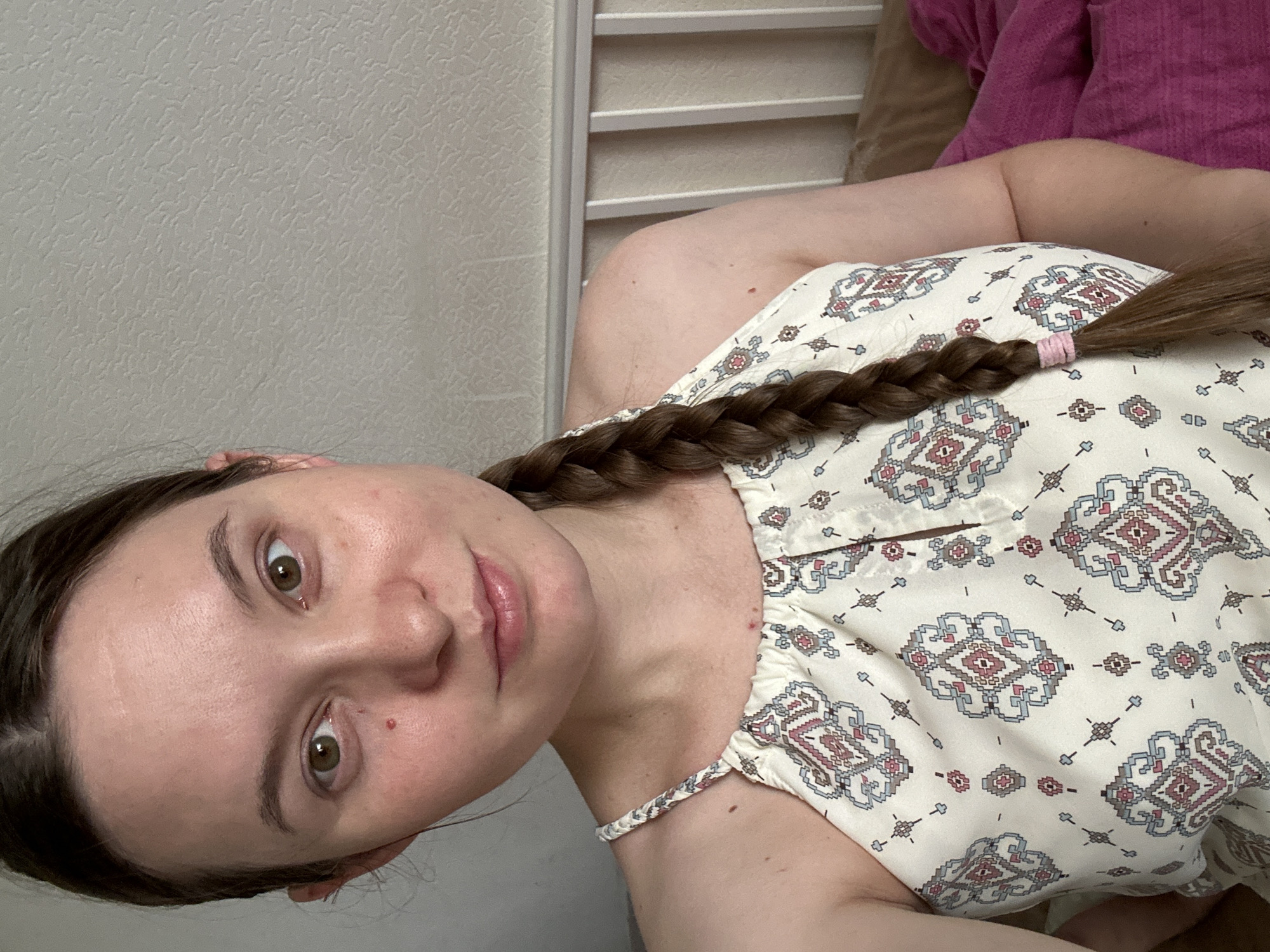 A young woman with brown hair braided down her left side is looking towards the camera in a close-up shot. She is wearing a white top with a light pink and blue geometric pattern. Her skin appears fair, and she has light brown eyes. The background suggests an indoor setting, possibly a bedroom, with a textured wall and a white headboard. There is no indication of the city of Noviy, Russia, within the image.