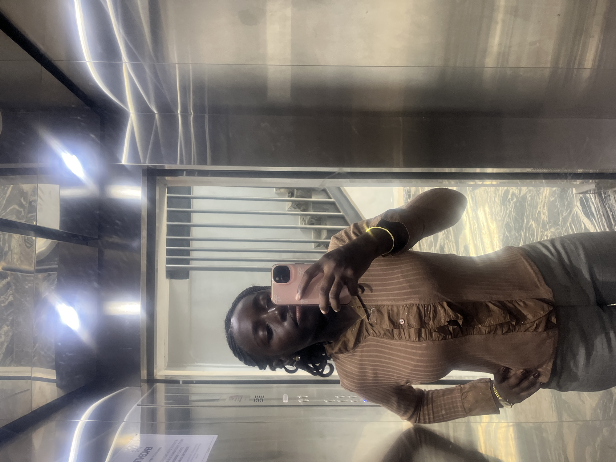 In the city of Karum, Nigeria, a young Black woman stands in an elevator, taking a selfie with her pink smartphone. She has dark skin, braided hair pulled back from her face, and is wearing a brown, pinstriped, ruffled blouse and grey pants. She has on gold bracelets on both wrists and a delicate gold necklace. The elevator walls are made of polished metal, reflecting the bright overhead lights and a staircase visible through an open doorway. A white sign with text is affixed to the left wall of the elevator. The floor of the elevator appears to be a patterned grey.