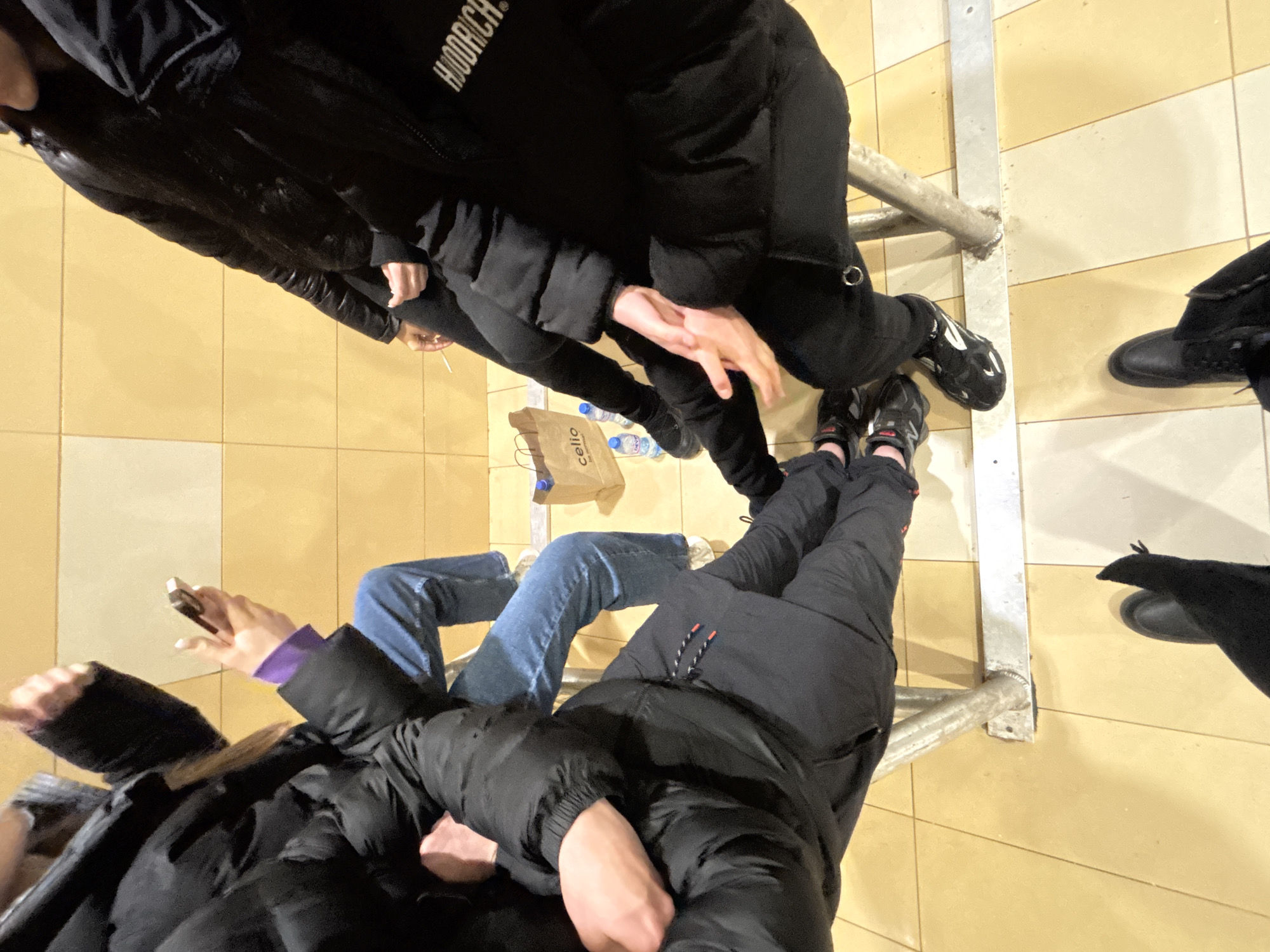 A high-angle shot shows a group of people standing on a tiled floor, possibly in a public area. The tile pattern is a grid of alternating light beige and cream squares. The scene features at least five individuals, identified by their legs and clothing. A metal railing runs diagonally across the frame, separating the group. Clothing consists of dark puffer jackets and blue jeans. A 'celio' paper bag and water bottles are on the floor near the railing. The location is potentially Sofia, Bulgaria.