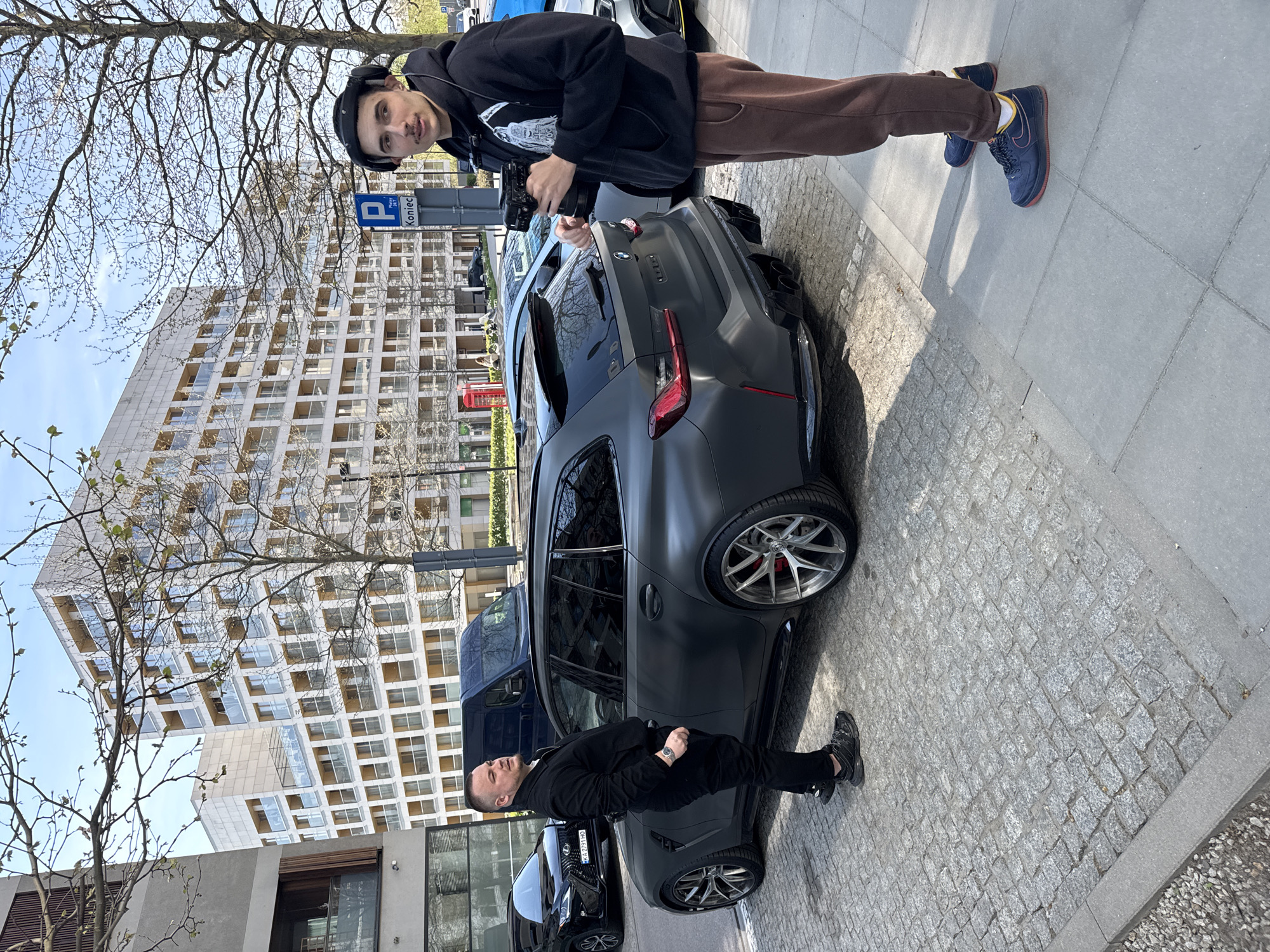The image shows two men next to a dark gray BMW M3 Touring. The man on the right is wearing a black hoodie, brown sweatpants, and blue sneakers, and he is holding a camera. The man on the left is wearing a black suit. Behind the car, there is a black Lexus SUV. In the background, there is a modern building with many windows. The street is paved with cobblestones. A parking sign is visible near the man on the right. Based on the architecture and street signage, the location is likely Warsaw, Poland.