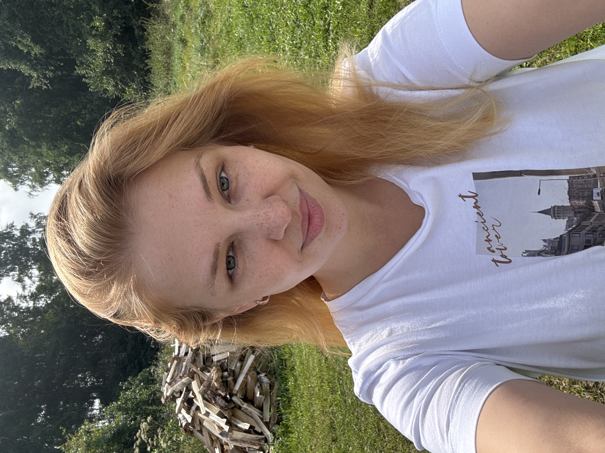 A close-up, selfie-style shot features a young woman with blonde hair and freckles. She is wearing a white t-shirt with a graphic design on it. The background shows a pile of chopped wood, green grass, and trees, suggesting an outdoor, rural setting. The image is taken in Cherkasovo, Russia.