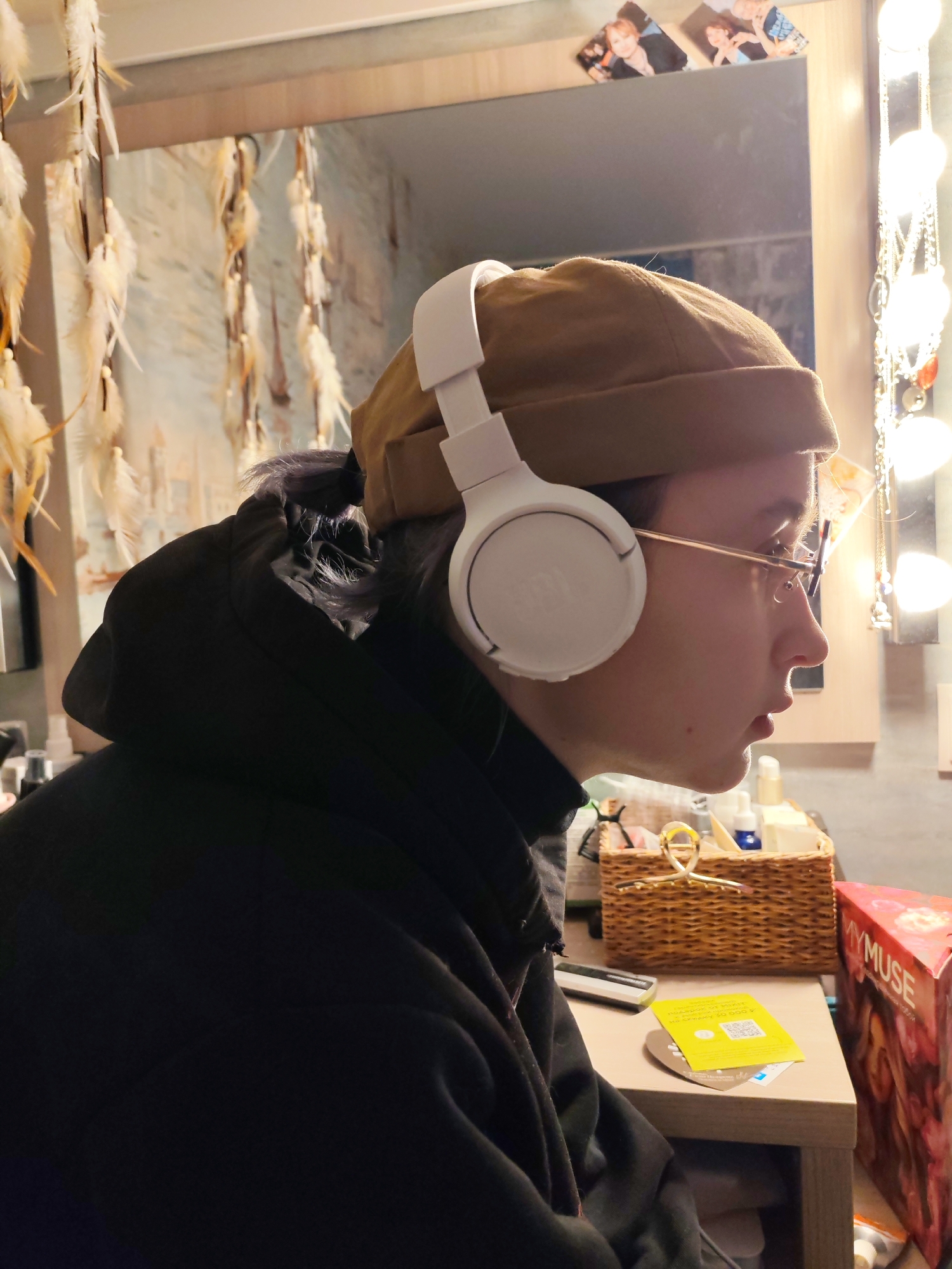 An indoor scene in Chaikovsky, Russia, depicts an individual in profile, facing right, positioned in front of a vanity mirror. The person wears a brown beanie, white JBL over-ear headphones, gold-rimmed glasses, and a dark hooded garment. Greyish-purple hair is visible beneath the beanie near the neck. Their mouth is slightly agape, indicating focused attention.
The background features a large rectangular mirror mounted on a light wooden surface. Two small photographs of young men are affixed to the surface above the mirror. A vertical string of illuminated bulbs, characteristic of vanity lighting, runs along the right edge of the mirror. To the left, several decorative elements composed of light-colored feathers or fronds are suspended.
A light-colored wooden desk or shelf occupies the lower right portion of the frame. Arranged on this surface are: a small dark rectangular electronic device, a yellow card displaying Cyrillic text (including "ПОДАРОК ОТ ХОКЕЙНОГО КЛУБА..." and a QR code), a partially visible blue and white credit/debit card, and a woven wicker basket. The basket contains various small bottles and containers, including a blue dropper bottle. A gold-colored, loop-shaped hair clip rests on the front rim of the basket. A large red and pink box, partially labeled "MY MUSE," is visible on the far right of the desk.