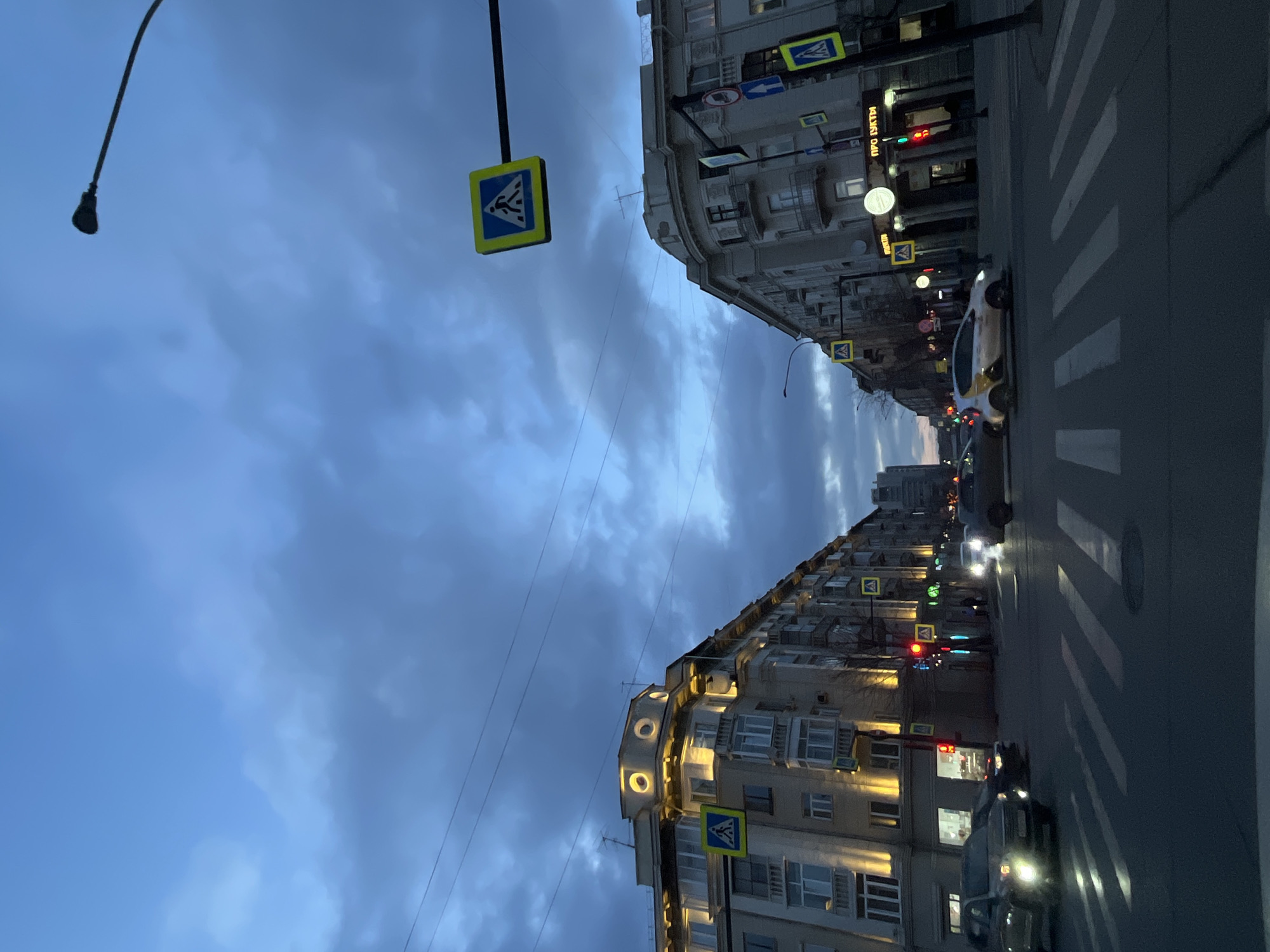 The image captures an urban street scene at dusk or early evening, taken from a low angle and rotated 90 degrees clockwise. The sky is largely overcast, displaying various shades of blue and gray clouds.
Multi-story buildings line both sides of the street, featuring numerous windows, some balconies, and architectural details. Many of the buildings' windows and ground-floor establishments are illuminated, casting a warm glow onto the street. A paved road with a clearly marked zebra pedestrian crossing runs through the lower and central part of the frame.
Several vehicles are present on the road, with some cars showing illuminated headlights, indicating the dim light conditions. A white car with yellow markings is prominently positioned near the crosswalk on the right side. The street is equipped with multiple road signs, including several distinctive yellow square signs with blue triangles and white figures, signaling pedestrian crossings. Other regulatory and directional signs, along with traffic lights (showing both red and green signals), are also visible. A dark, curved lamppost extends into the upper left corner of the sky. Utility wires traverse the sky between the buildings.
On one of the illuminated building facades, a neon sign displays partial Cyrillic text that reads "ПРО БУТЫ". The overall setting depicts a typical city street, bustling with activity, consistent with an urban environment like Krasnoyarsk, Russia. No people are distinctly visible in the scene.