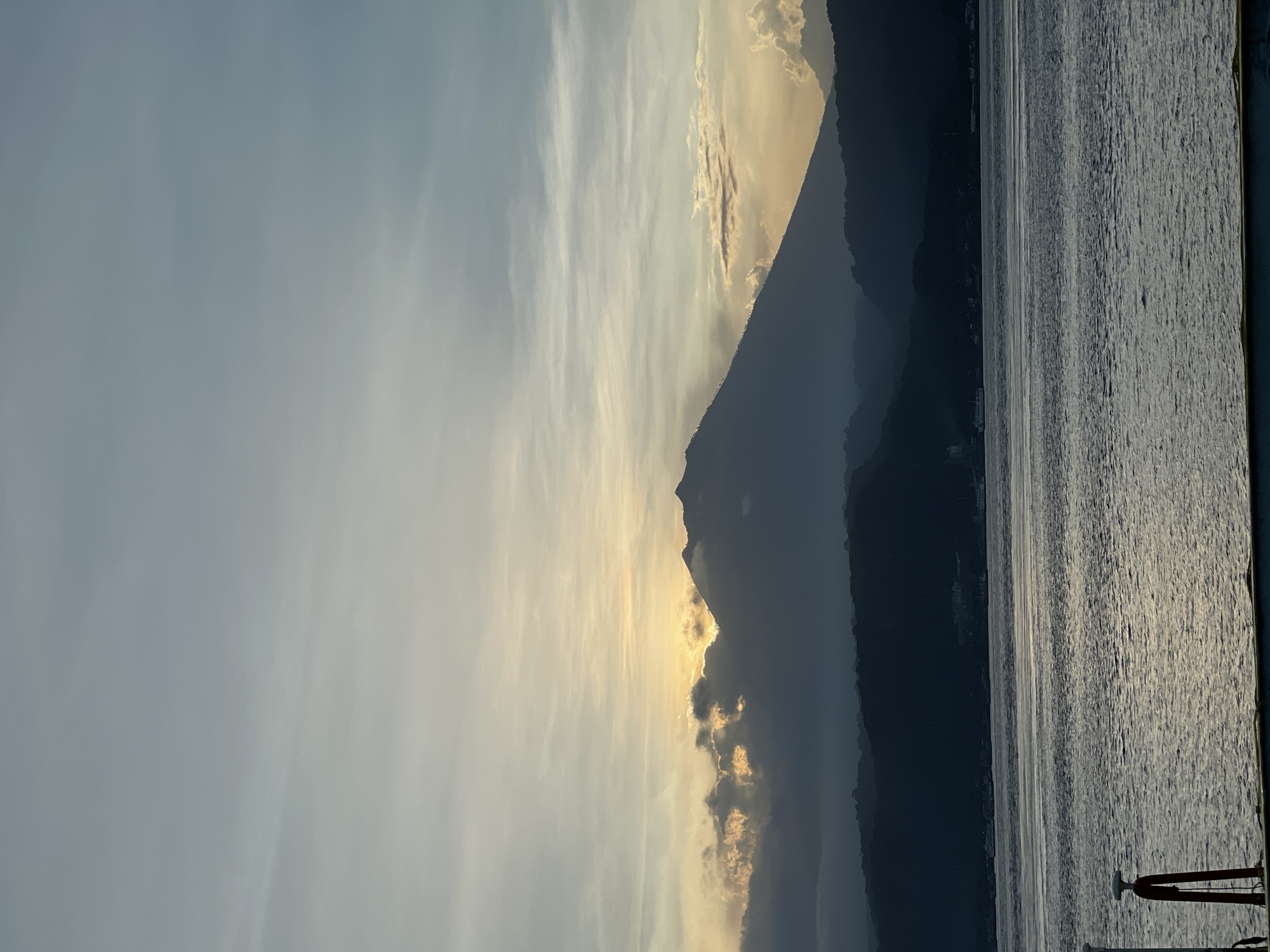 The image depicts a mountainous landscape adjacent to a body of water, likely a lake or sea. The sky is partly cloudy with a golden light source, possibly the sun, peeking through the clouds and creating a dramatic effect. A dark, silhouetted mountain range runs along the right side of the image. The water surface is textured, reflecting the light. A portion of what appears to be a boat or pier is visible on the far right, evidenced by a vertical pole and a red object at the bottom. No people are visible in the frame. The location is unspecified.
