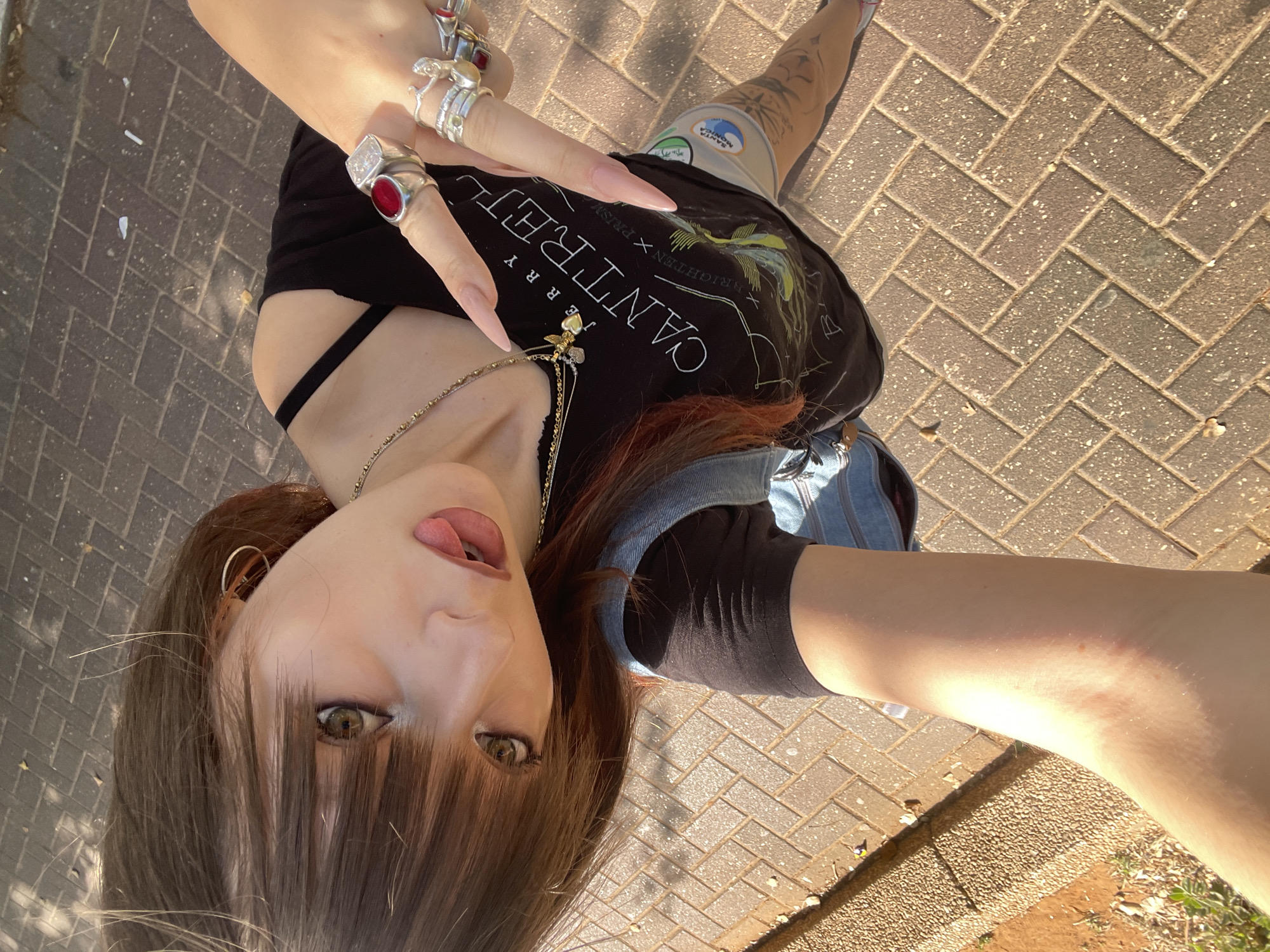 A young woman with auburn hair and bangs is taking a selfie outdoors on a paved walkway. She is wearing a black t-shirt with a graphic print and a denim vest. She has multiple rings on her fingers, including one with a large red stone and another with a clear rectangular stone. Her nails are long and light pink. The background shows a brick pathway and some greenery. The lighting suggests it is daytime. The provided image hints indicate the location is Raanana, Israel.