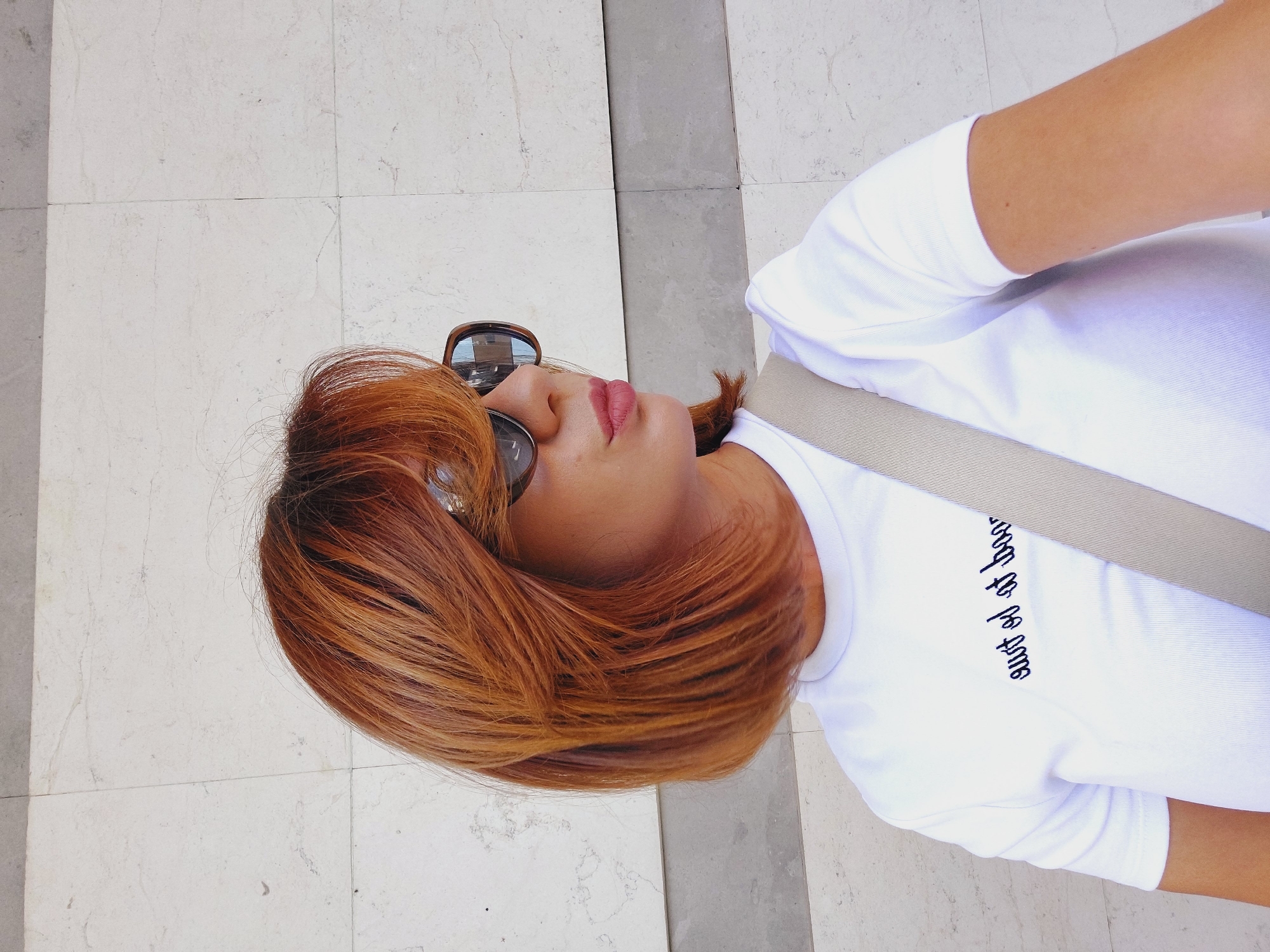 A woman with auburn hair and sunglasses is posing for a selfie in front of a marble wall in Sofia, Bulgaria. She is wearing a white t-shirt with a printed message in dark blue cursive letters. A beige strap is visible across her shoulder, likely from a bag. The wall behind her is made of large marble tiles with a faint grey stripe running horizontally across the frame. The lighting suggests it is daytime.