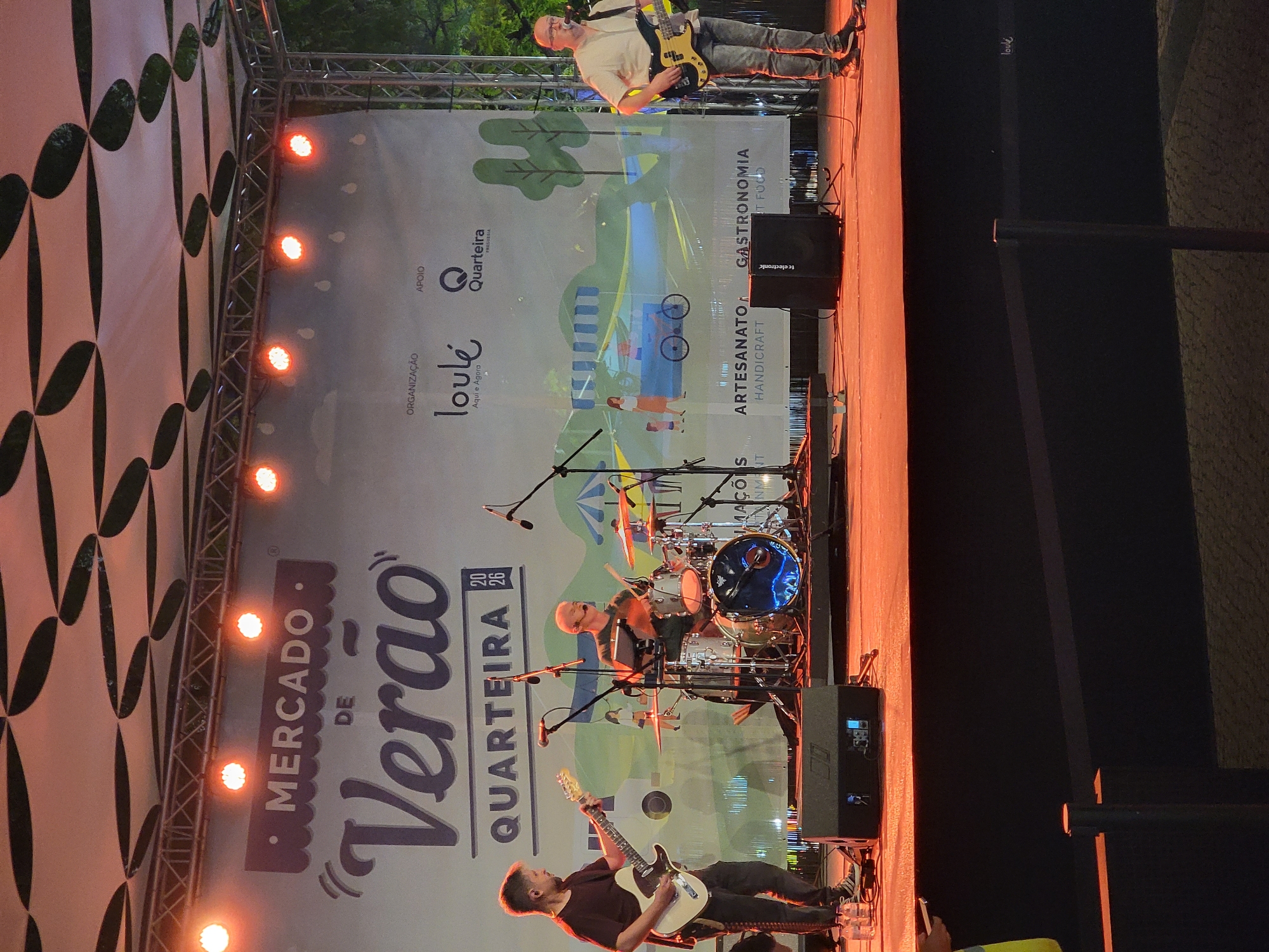 The image depicts a live music performance taking place on an outdoor stage at the "Mercado de Verão" (Summer Market) in Quarteira, Portugal. The backdrop is a large banner with the event's title and logos. Three male musicians are visible on stage. On the left, a guitarist wearing a dark t-shirt and dark pants plays a white electric guitar. In the center, a drummer in a green t-shirt is actively playing a drum set. On the right, a bassist wearing a light-colored short-sleeved shirt and gray jeans sings and plays a dark electric bass guitar. The stage is illuminated by warm-toned lights, and lighting trusses are visible above the stage. Several microphones and audio equipment, including speakers and monitors, are arranged on the stage. The background banner features cartoonish illustrations of market stalls and trees, with "ORGANIZAÇÃO LOULÉ AQUI E AGORA" and "APOIO QUARTEIRA PROCURESIA" text. The overall environment suggests a festive evening event in Quarteira.