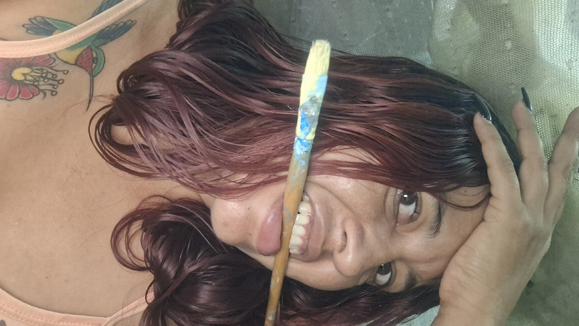 A woman with dark, wavy hair, partially wet and dyed reddish-brown, is shown from the chest up. She is holding a paintbrush horizontally in her mouth, with the bristles pointing to the left and stained with blue and yellow paint. Her right hand is placed on her forehead, with black painted fingernails visible. The woman has dark eyes and a light complexion. She is wearing a light-colored tank top, and a tattoo of a hummingbird and a flower is visible on her left shoulder and upper chest. The background appears to be a textured, light-colored surface, possibly a wall or a curtain. The image appears to be taken indoors. This scene is not specifically tied to a location in Vallecito, Venezuela.