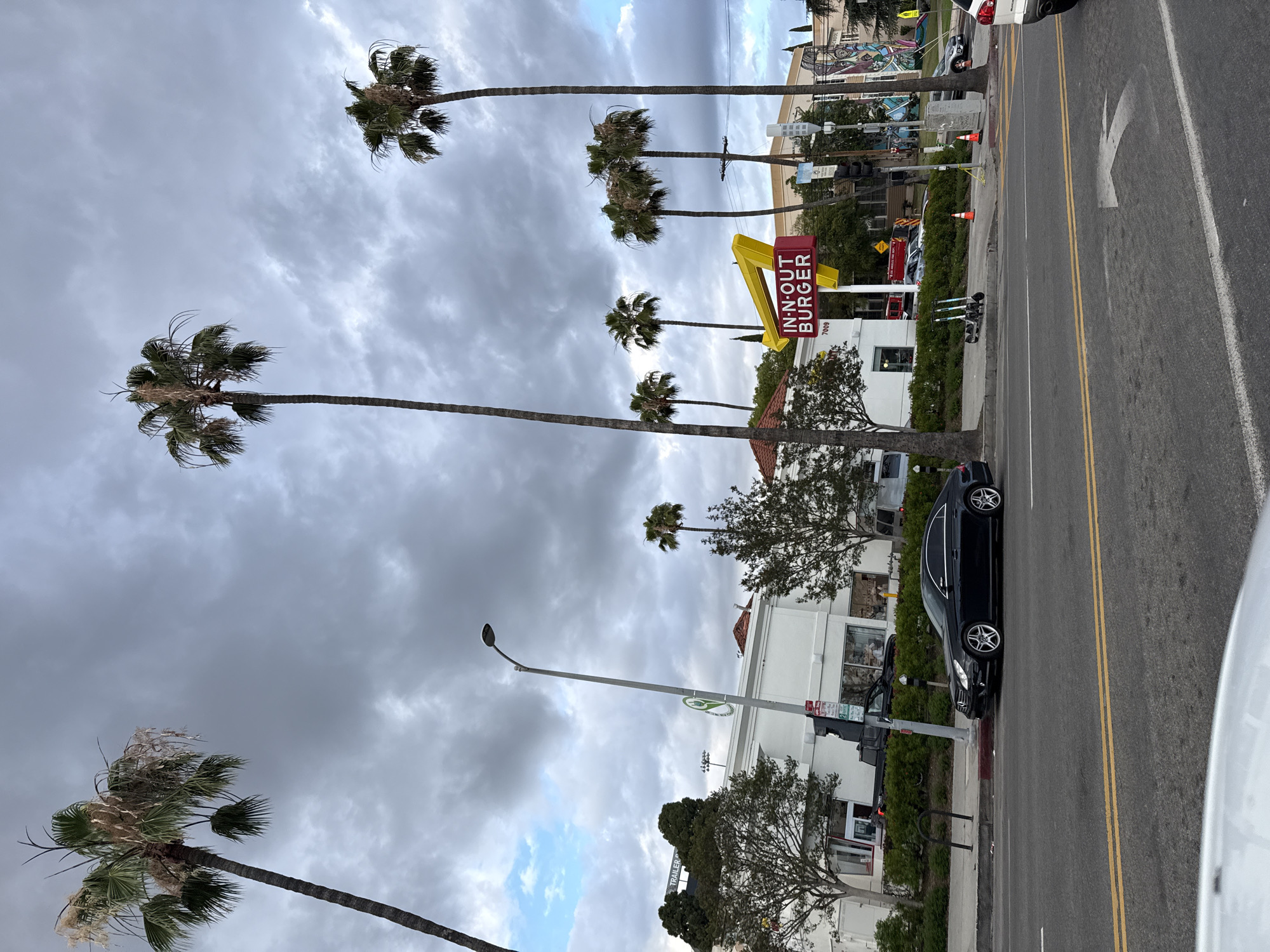 The image captures an urban streetscape in West Hollywood, California. Palm trees line the street under a cloudy sky. An In-N-Out Burger restaurant is visible, with its distinctive yellow and red signage. A black sedan is parked along the curb. The street has lane markings, and a street lamp is present. A mural or artwork is visible in the background. Other buildings and some vehicles are also part of the scene.