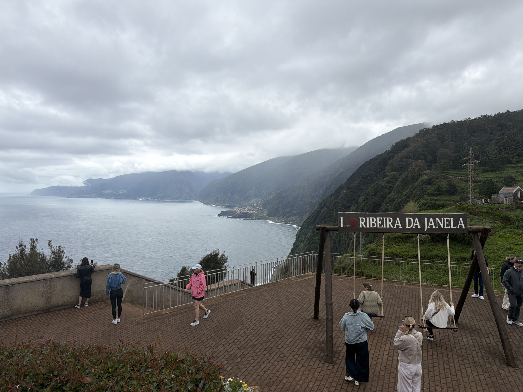 The image shows a view of Ribeira da Janela, a coastal area in Porto Moniz, Portugal. People are present on a paved area with a lookout point. Some individuals are taking photos of the landscape and others are interacting with a swing set displaying the words "I <3 LOVE> Ribeira da Janela." The landscape includes a coastline with cliffs, mountains covered in vegetation, and the ocean. The sky is overcast with clouds.