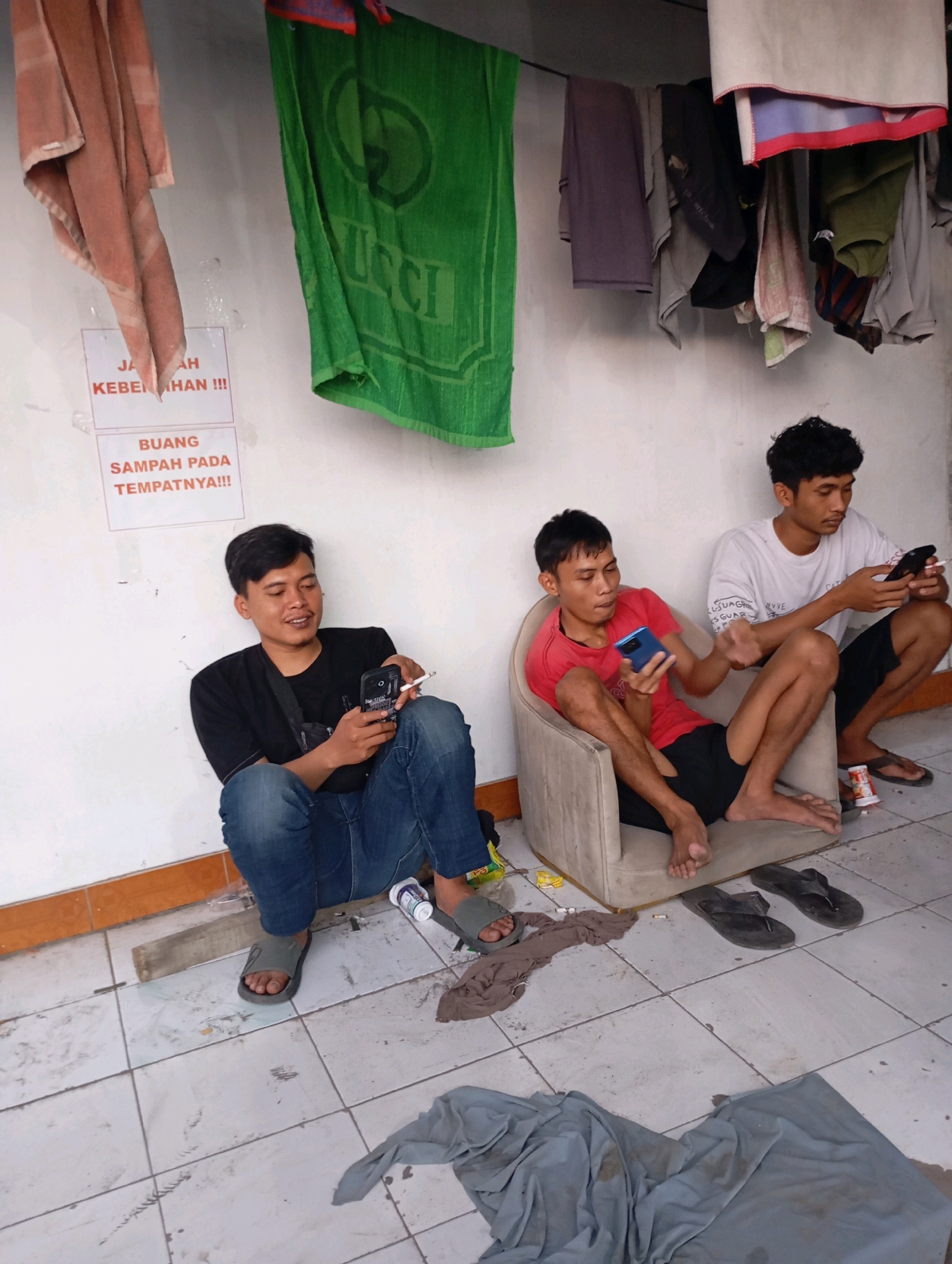 The image depicts an indoor scene, likely in a dormitory or communal living space in Tasikmalaya, Indonesia, evidenced by the Indonesian text on the signs. Three young men are present. On the left, a man in a black t-shirt and blue jeans is seated on the floor, holding a cigarette and a smartphone. In the center, another man in a red t-shirt and black shorts sits in a beige armchair, also holding a smartphone. To the right, a third man in a white t-shirt and dark shorts is crouched on the floor, looking at his smartphone. Laundry hangs on a clothesline above them against a white wall. Two signs are posted on the wall; one reads "JAGA KEBERSIHAN!!!" (Maintain Cleanliness!!!) and the other "BUANG SAMPAH PADA TEMPATNYA!!!" (Dispose of Trash in its Place!!!). The floor is tiled and scattered with clothing and debris.