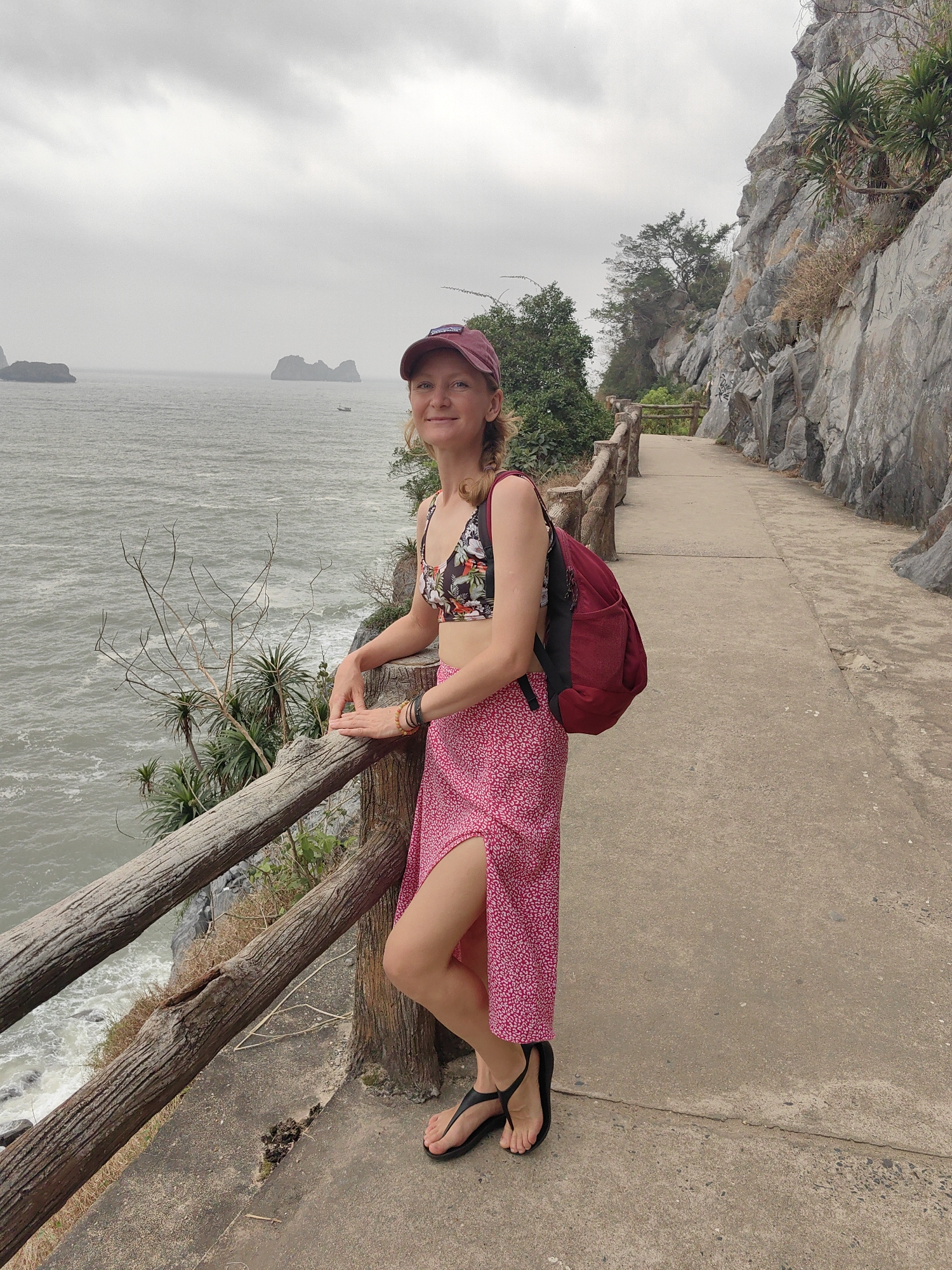 A woman is standing on a concrete path next to a wooden railing in a scenic outdoor setting. She is wearing a red cap, a floral print bikini top, a long pink skirt with a side slit, black sandals, and a red backpack. The path is bordered on one side by a rocky cliff face with some vegetation growing on it. On the other side of the railing, there is a view of the ocean with islands in the distance under an overcast sky. The location is Cát Bà Island in the city of Hải Phòng, Vietnam.