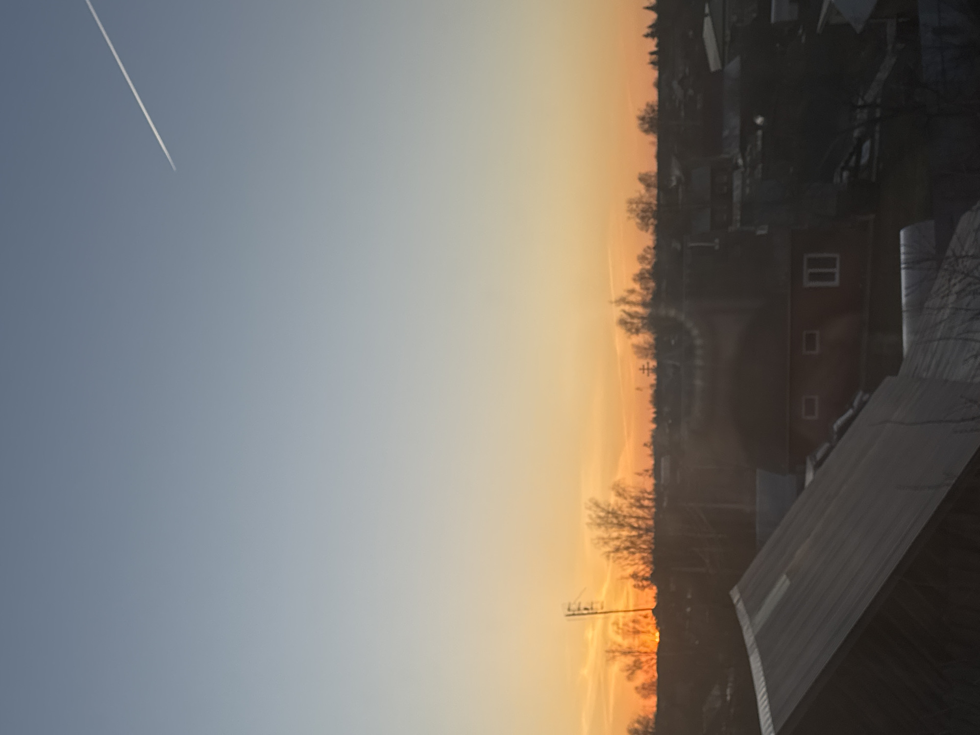 The image shows a landscape at dawn or dusk in Фоки, Russia. The sky transitions from blue to orange, with a contrail visible in the blue part. In the foreground, there's the corner of a building with a metallic roof, possibly a house or industrial structure. The middle ground contains trees without leaves silhouetted against the orange sky. Further back, other buildings and urban structures can be seen, some illuminated by the low sun.