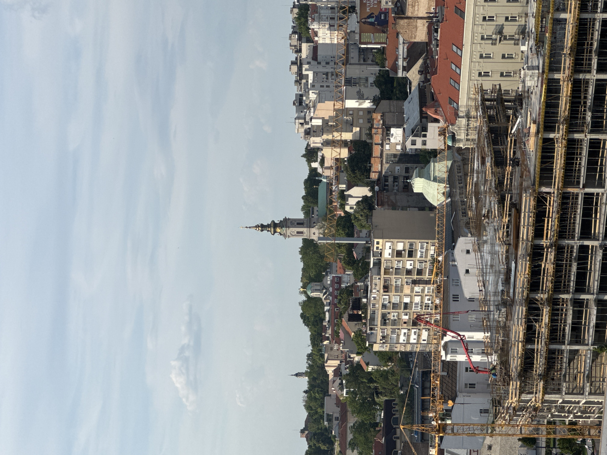 This image shows a cityscape in Belgrade, Serbia, featuring a view that includes the city's architecture, sky, and construction activities. In the mid-ground, there are residential and commercial buildings of varying heights and architectural styles. A church steeple is prominent among the buildings, marking a historic or religious landmark. A large construction site dominates the right side of the image, featuring a building under construction. The construction site includes scaffolding and construction equipment, indicating ongoing development. The sky is visible in the background, indicating the weather and providing context for the environment.