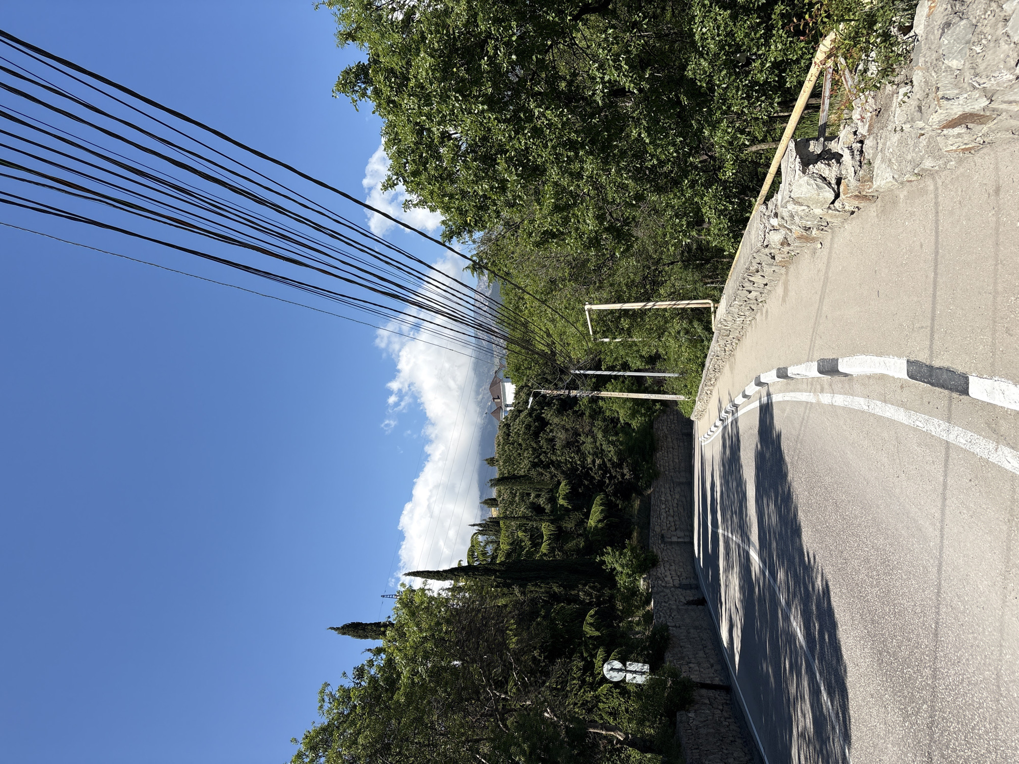 The image depicts a view from a winding road in the city of Ялта, Ukraine. The road surface is asphalt, marked with a white lane divider and black and white striped barriers along the edge. A stone wall borders one side of the road, with trees and vegetation lining the other side. Overhead, a large number of black power lines stretch across the sky, contrasting with the clear blue sky and scattered white clouds. In the distance, a building is visible among the trees, situated on a hillside.