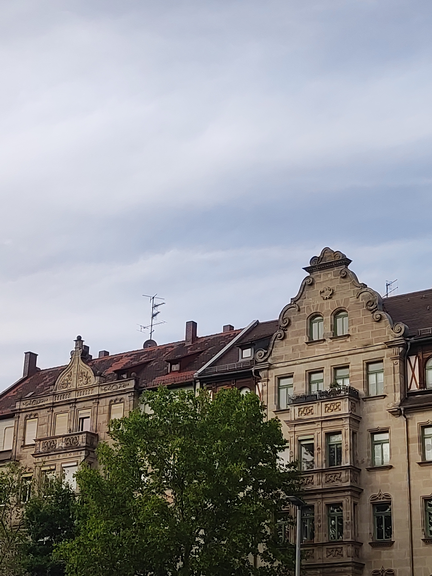 This is a vertical, eye-level, medium shot of a building's facade in Fürth, Germany. The building appears to be an old residential structure, with ornate stonework and red-tiled roofs. The facade is made of light-colored stone with decorative carvings around the windows and gables. There are multiple windows with green frames, some with closed shutters. A balcony with intricate railings is visible on the second floor. A large, leafy green tree partially obstructs the lower part of the building on the left side. The sky is overcast with pale blue and white clouds. An antenna is visible on the roof in the upper left portion of the image.