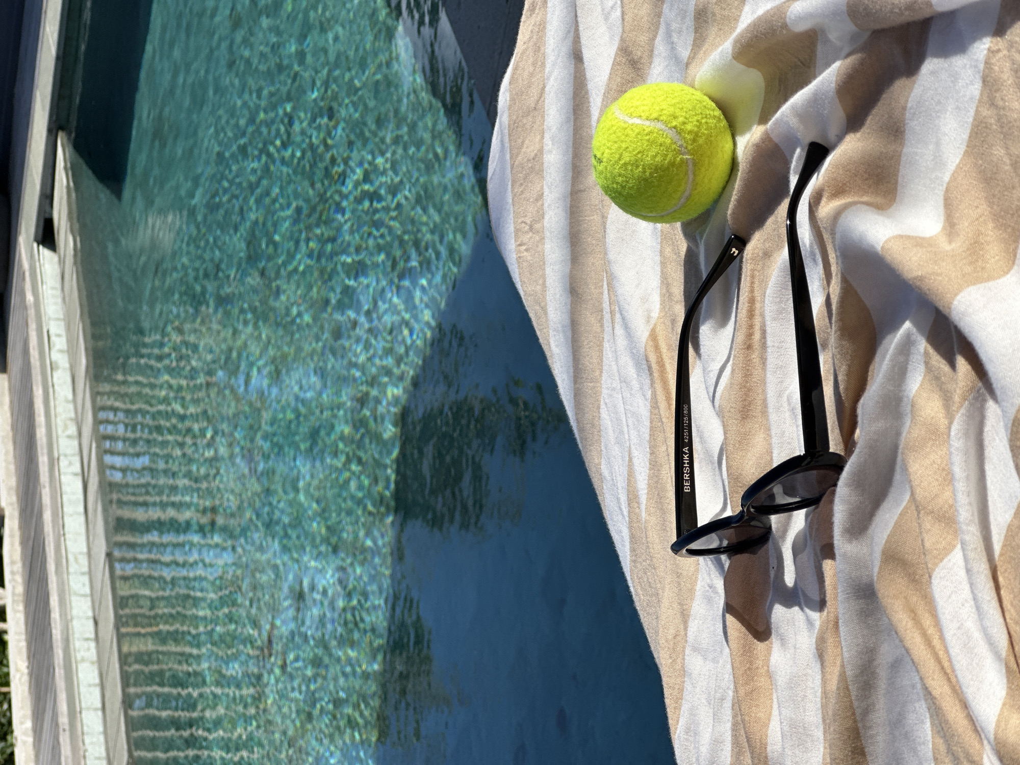 A pair of black sunglasses rests on a striped towel next to a tennis ball by the edge of a swimming pool in Kuta Selatan, Indonesia. The pool water is a clear turquoise, reflecting the sunlight and the surrounding environment. The towel has alternating stripes of white and a light beige color. The tennis ball is a vibrant yellow-green, a stark contrast to the towel and sunglasses. The poolside is made of wooden planks.