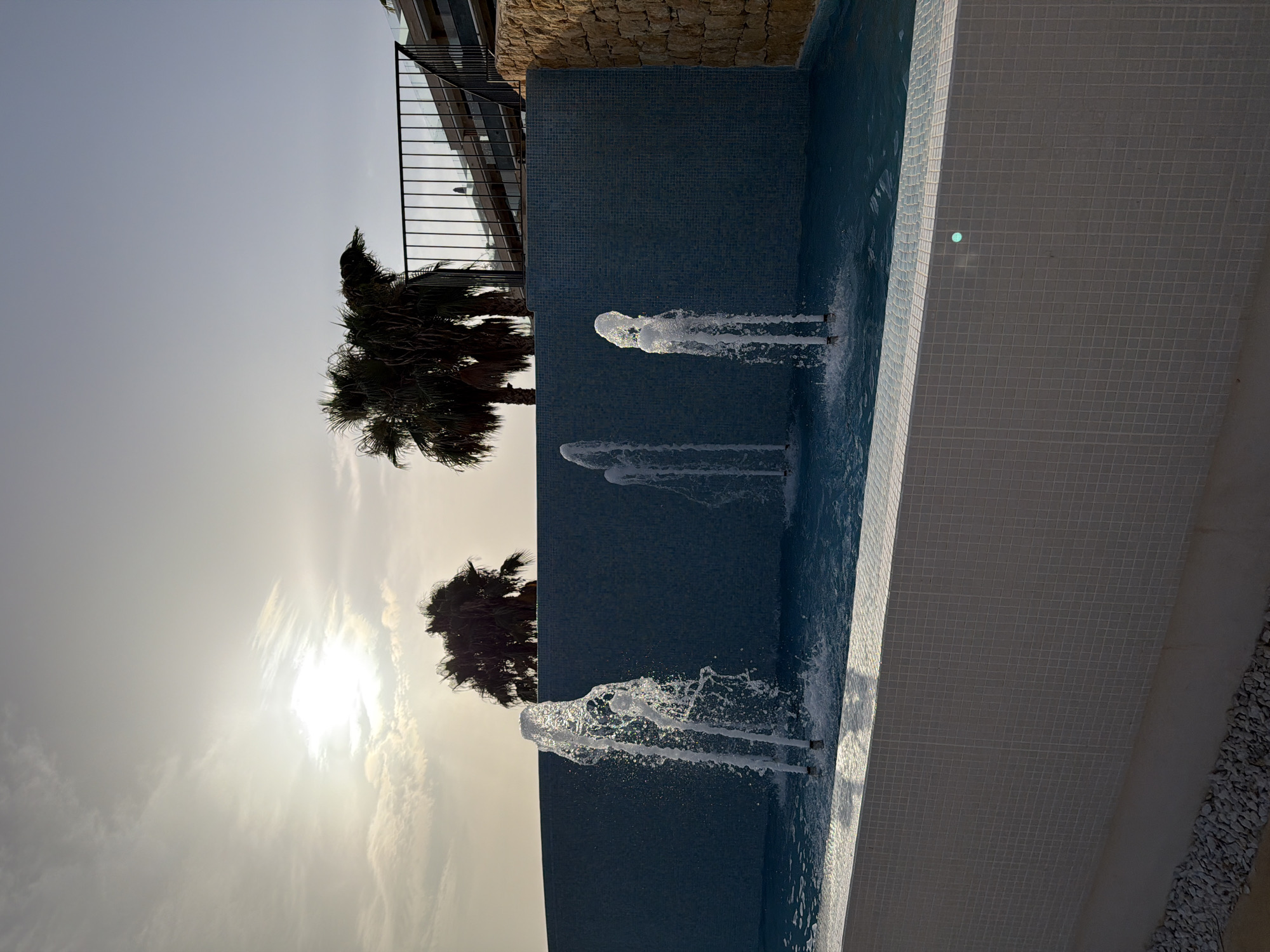 This is a vertical, medium shot of a swimming pool area with fountains in Finestrat, Spain. The scene is bathed in the warm, hazy light of sunset. The pool's edge is lined with small white tiles, and a dark blue tiled wall forms the backdrop for several water fountains that are actively spraying water into the pool. The water from the fountains is captured in mid-air, creating a dynamic and refreshing effect.
In the background, silhouetted against the bright sky, are two palm trees, their fronds dark and feathery. Beyond the pool and palm trees, a modern building with dark railings is visible, and a stone-tiled wall adds texture to the upper part of the frame. The sky is a soft gradient of pale blue and white, with wispy clouds catching the golden light of the setting sun. The overall mood is serene and relaxing, suggesting a warm evening in a resort setting. There is no visible text in the image.