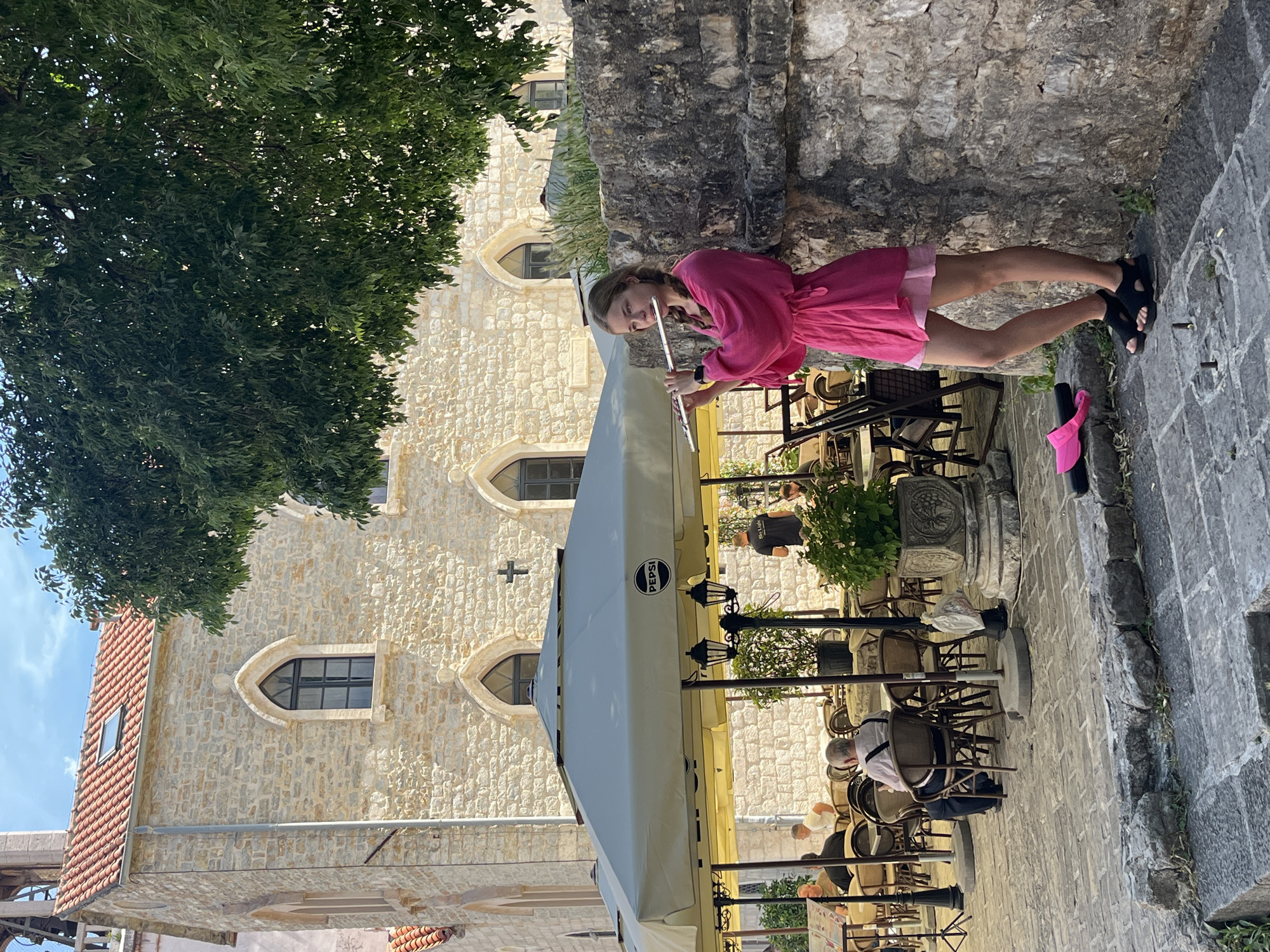 A woman in a pink dress stands on a stone street in the city of Budva, Montenegro. She is playing a silver flute. Behind her is a stone wall and a stone building with arched windows. In front of her is a white awning, tables and chairs, and people sitting at the tables. A pink visor is on the street near her feet. A tree is on the left side of the image.