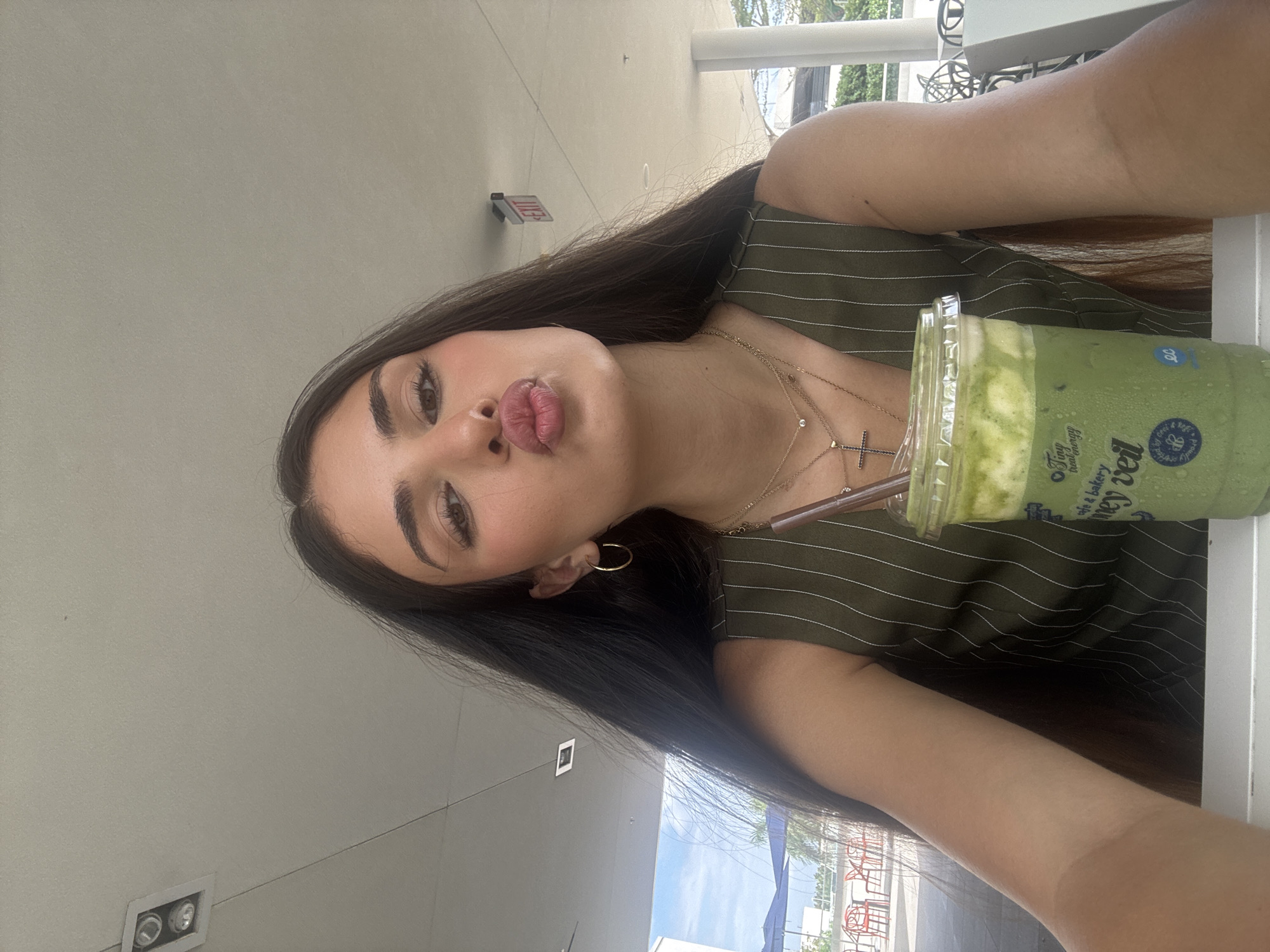 A young woman with long, dark hair and a green pinstripe top is seated outdoors in Hallandale Beach, Florida, USA. She is holding a clear plastic cup containing a green beverage with a white foamy top and a brown straw. The cup has blue text and graphics on it. She is puckering her lips and looking towards the camera. She is wearing two gold necklaces, one with a small diamond and the other with a cross pendant. She also has a gold hoop earring in her left ear. The background shows an outdoor seating area with red chairs and tables, blue umbrellas, and buildings. The ceiling above is light grey.
