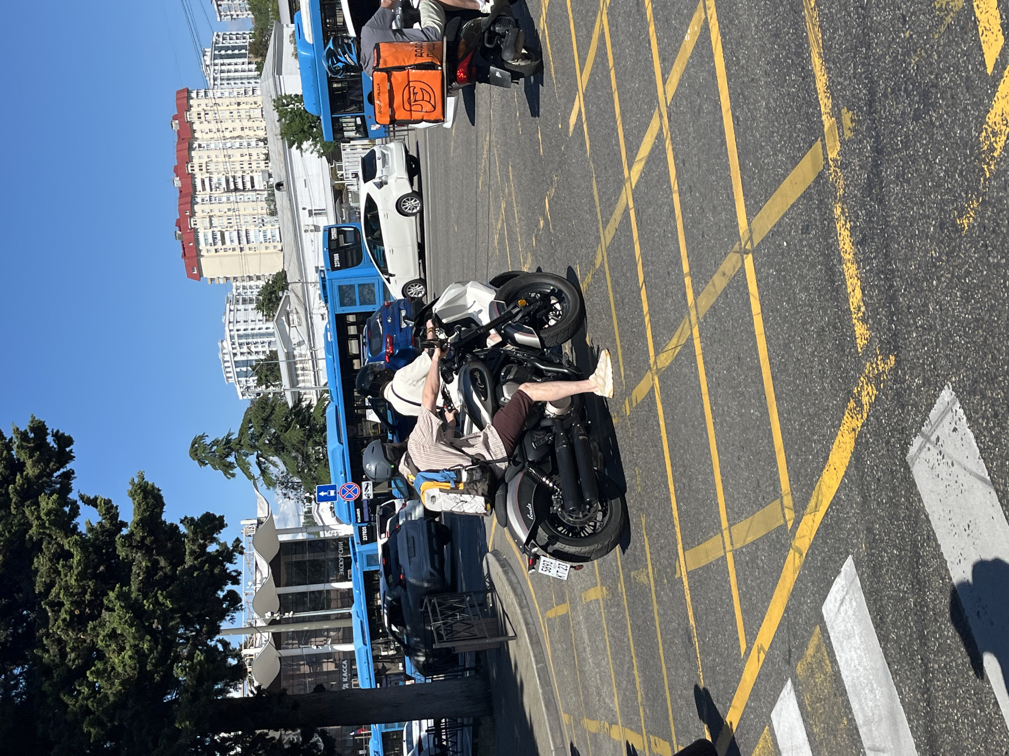 In Sochi, Russia, on a sunny day, a motorcycle carrying two individuals passes through a street intersection marked with yellow grid lines and a pedestrian crossing. The motorcycle, a dark cruiser-style bike, is ridden by a person wearing a helmet and a striped shirt, with a backpack on their back. The passenger behind them is also wearing a helmet and a light-colored shirt. To the right, a scooter with a large orange delivery box, labeled "DOÐO PIZZA," is being ridden by a person wearing a helmet. In the background, blue buses, cars, and apartment buildings are visible under a clear blue sky. The scene appears to be a typical urban street in Sochi, with traffic and pedestrians navigating the city.