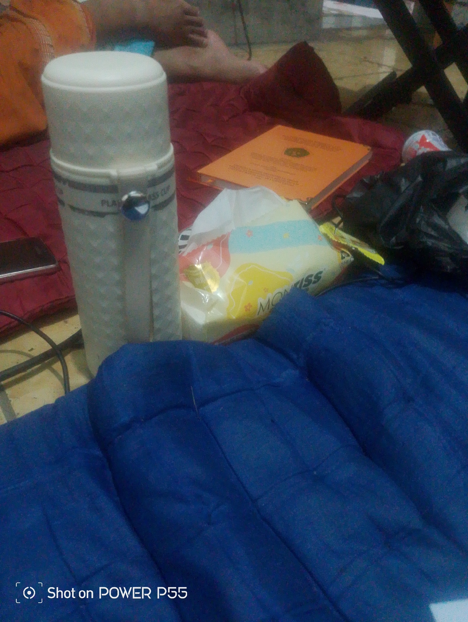 In a dimly lit room, possibly a bedroom in Lebaksiu, Indonesia, a white thermos with a textured surface stands upright on a red cloth. A blue circular object is affixed to the side of the thermos. Beside the thermos is a packet of tissues with a yellow and blue design, and the word "MONASS" is partially visible. To the left of the thermos, a dark rectangular object, likely a smartphone, rests on the red cloth. Behind these items, a person's legs are visible, one with a patterned orange garment on the thigh. The scene is laid out on a red fabric, with a portion of a dark blue quilted surface occupying the foreground.