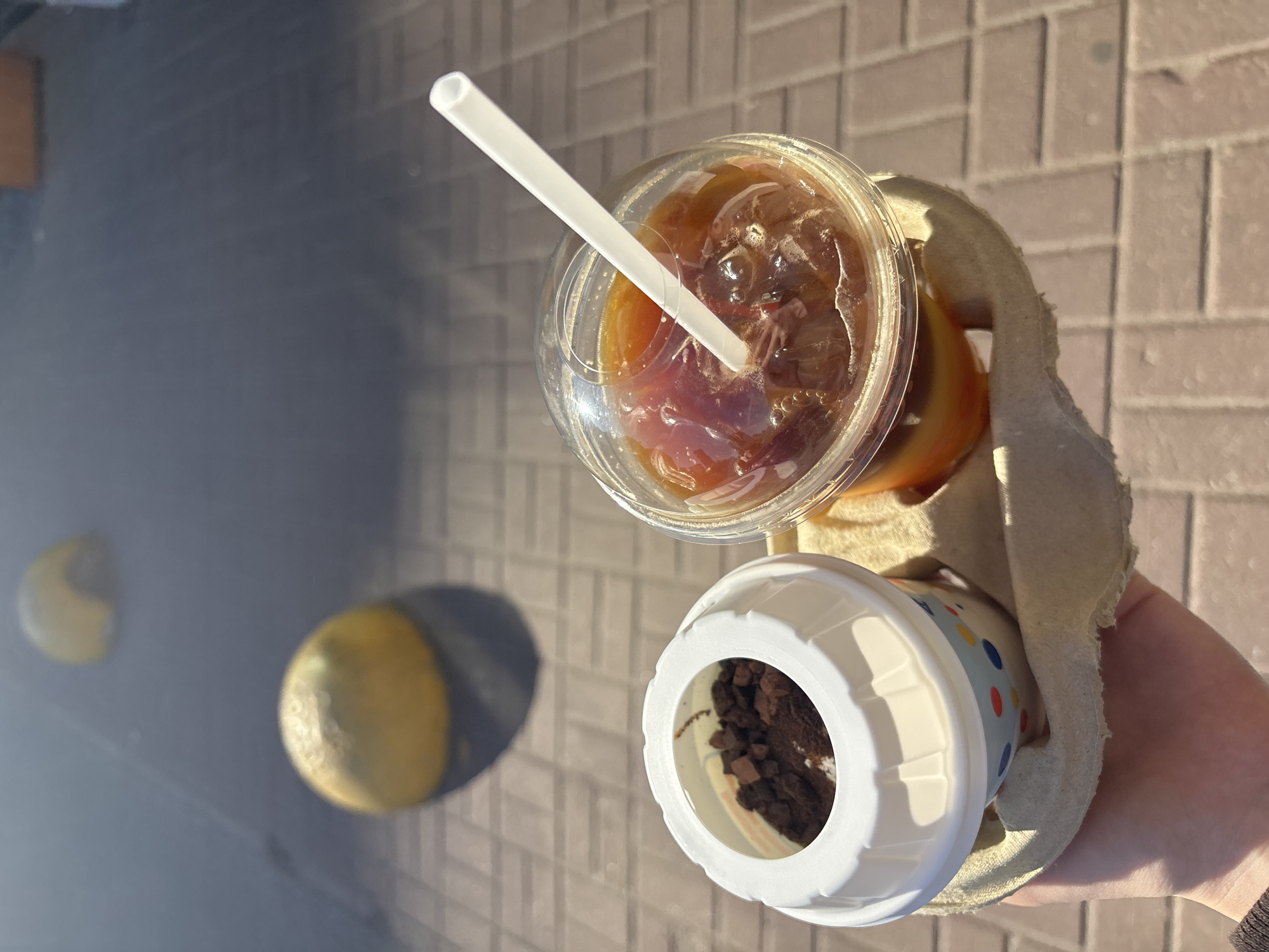 The image shows a hand holding a cardboard carrier with two beverages: an iced coffee with a white straw and a small cup filled with what appears to be chocolate crumbles or a dessert. The background features a walkway or sidewalk paved with rectangular stones and two rounded bollards, painted yellow and black, partially visible. The location is implied to be in Vsevolozhsk, Russia. The scene is outdoors, with sunlight creating shadows and highlights.