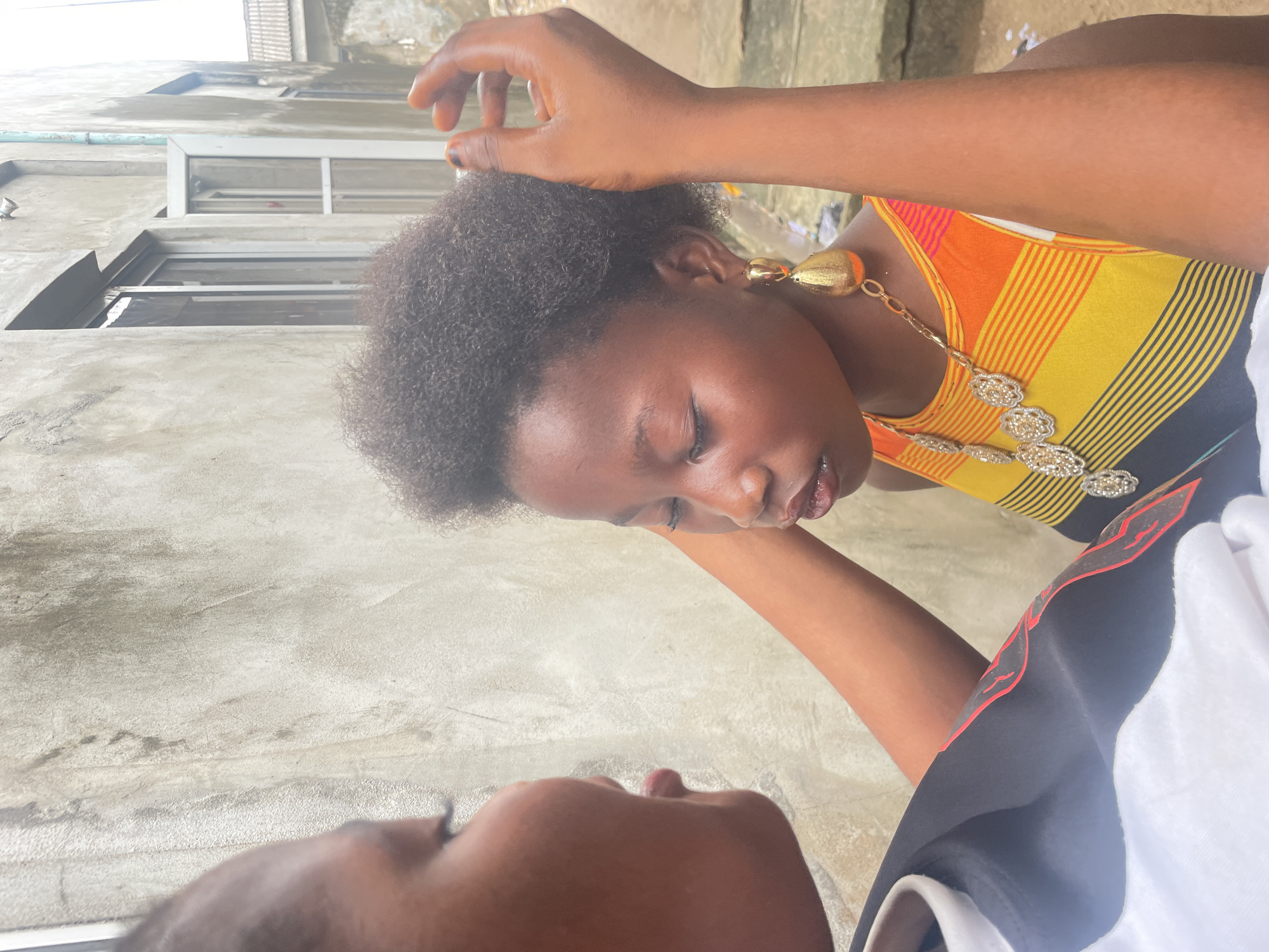 The image, rotated onto its side, captures a close-up interaction between two individuals in what appears to be an outdoor or semi-outdoor setting in Badagry, Nigeria, during the daytime.
In the mid-ground, a person with dark skin and short, curly black hair is the primary subject. They are wearing a brightly colored, vertically striped top in shades of orange, yellow, and black. A prominent, ornate golden necklace with multiple decorative elements adorns their neck, complemented by golden teardrop-shaped earrings. Their eyes are mostly closed or looking downwards, suggesting concentration or tenderness, and their lips are slightly parted. One of their hands, with dark nail polish, is raised and positioned near their own head or the other person's head, implying a gentle touch or gesture.
In the foreground, partially visible, is the top of another person's head and face, including their forehead, nose, and chin. This individual also has dark skin and is looking upwards towards the first person. They are wearing a white garment with black sections and subtle red detailing.
The background consists of a light gray, textured concrete wall, indicative of a building exterior. Several windows with frames are visible within this concrete structure. The bright, even illumination suggests it is daytime, with soft lighting preventing harsh shadows. The visible ground beneath them is also a light-colored, textured concrete surface. No discernible text is present in the image. The overall scene conveys an intimate and focused moment between the two individuals.