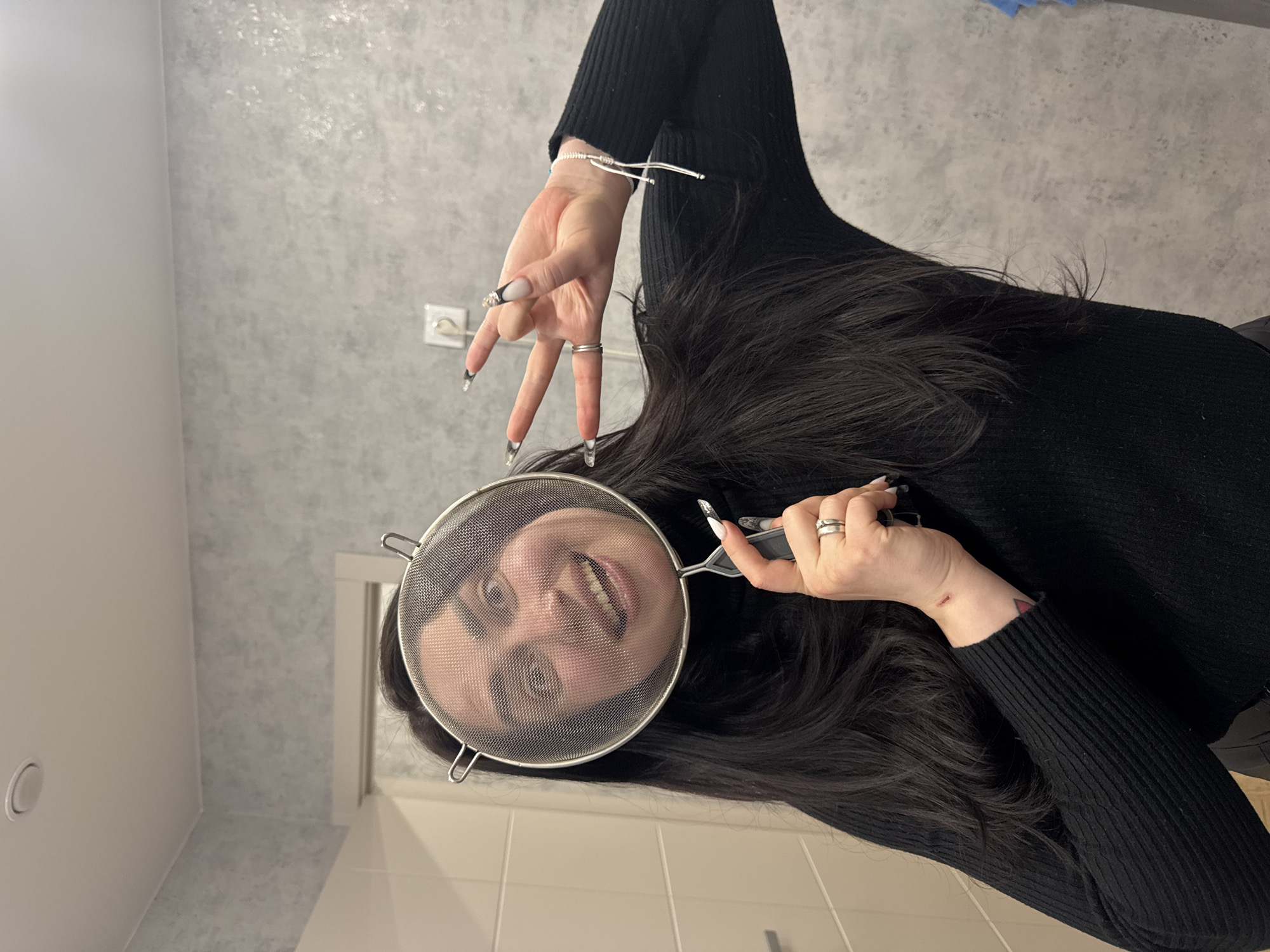 A playfully composed, vertically oriented photograph captures a person, likely a woman, indoors. She has long, dark hair that cascades around her shoulders and is wearing a black, ribbed, long-sleeved top.
The central activity shows her holding a round metal kitchen sieve or strainer directly in front of her face. Her eyes are visible through the mesh, and she is smiling broadly, revealing her teeth, indicating a lighthearted or humorous mood.
Her right hand (on the viewer's left) is raised and positioned to the side of her head, displaying long, pointed fingernails with a distinct black and white tip design. This hand is making a gesture with the index and middle fingers extended upwards, while the ring and pinky fingers are curled down. A silver bracelet is visible on her wrist, and a silver ring is on her ring finger. Her left hand (on the viewer's right) firmly grips the handle of the sieve.
The background consists of a textured, mottled grey wall, possibly with a stucco or concrete finish. A white electrical outlet is visible on this wall. The lower portion of the image shows a lighter, possibly tiled, beige or cream-colored wall or floor. A white ceiling with a circular fixture (possibly a light or smoke detector) is seen in the upper left corner. A white doorframe or corner is also visible on the right side of the frame. The lighting appears to be artificial, typical of an indoor setting. There is no visible text in the image.