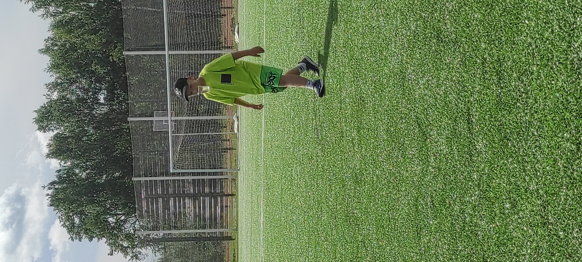 A young boy wearing a neon green t-shirt, black baseball cap, and green shorts walks on a green artificial turf field. He is holding a clear glass of water in his right hand, with the glass resting on his lower lip. The field appears to be a soccer field, with white lines visible on the turf and a soccer goal and net in the background. Behind the goal, there is a fence and then a line of trees under a cloudy sky. The location is identified as Noviy, Russia.