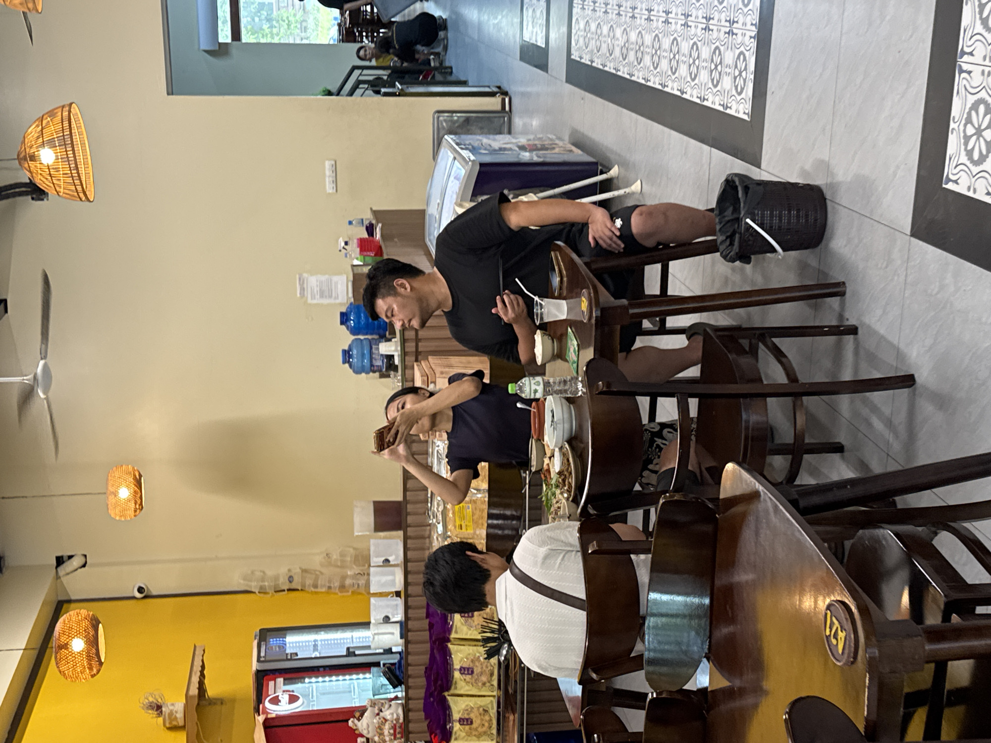 The image depicts an indoor dining area in a restaurant, likely in Hoa Lư Ward, Vietnam. In the foreground, a man wearing a black t-shirt and shorts sits at a wooden table, holding chopsticks and looking upwards. He is positioned to the right of a woman in a dark t-shirt, who is seated at the same table and is taking a photo with her phone. A white shirted man is seated to their left, facing away from the camera. The table is set with food, bowls, and drinks. Behind the table, a dark brown trash bin is visible on the floor. The environment features yellow and beige walls, with two rattan pendant lamps hanging from the ceiling. In the background, a refrigerator unit and shelves stocked with goods are visible, along with other patrons and a staircase leading to an upper level. The floor is tiled with a pattern of grey and white squares.