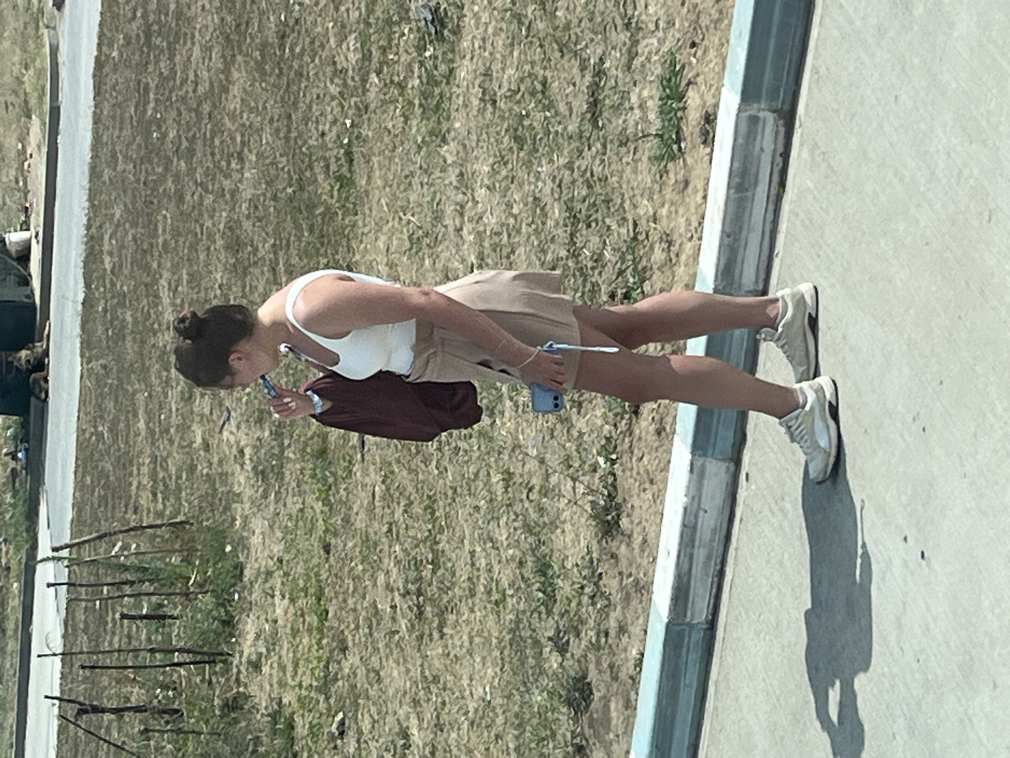 A young woman with her hair in a bun stands on a sidewalk, facing right. She is wearing a white tank top, a tan skirt, and white sneakers. She is holding a blue object, possibly a phone, in her right hand and a dark bag in her left. A faint shadow of the woman is cast on the ground in front of her. The background consists of dry grass and some wooden poles. In the distance, a road and some structures are visible. The scene appears to be in a semi-urban or rural area. The location is identified as Ovidiu, Romania.