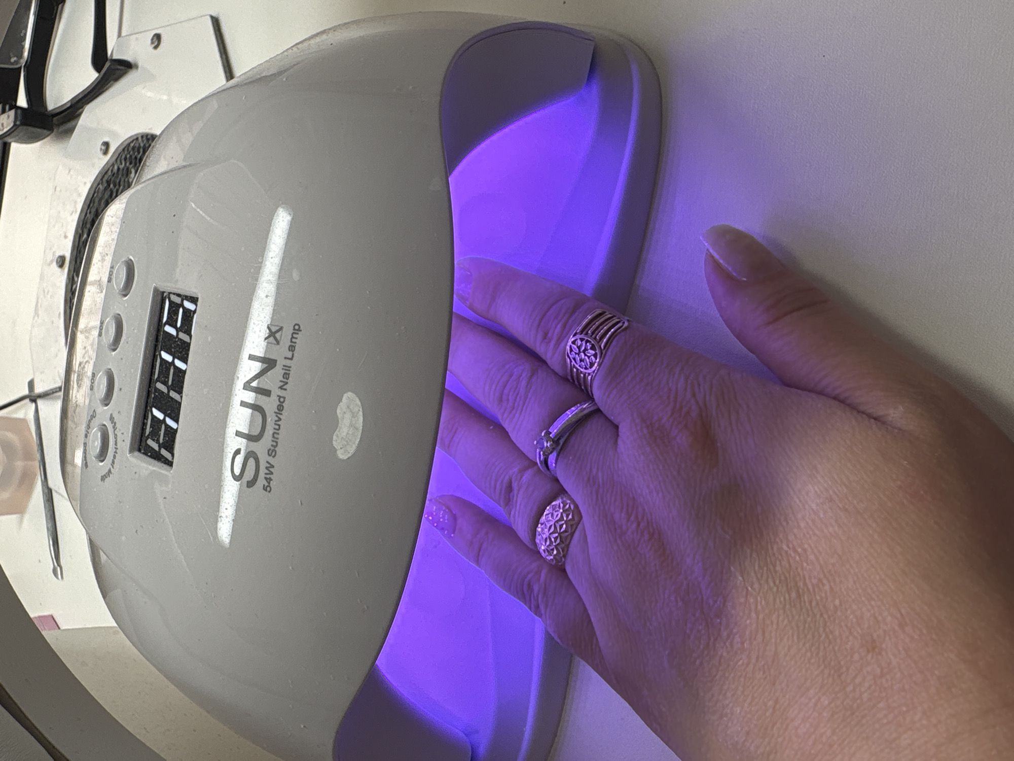 A human hand is placed inside a white SUNx 54W Sunuvled Nail Lamp. The lamp is emitting purple light. The hand has three rings on its fingers. The background is a light-colored surface. The specific location in the city of Noviy, Russia, cannot be determined from the image.