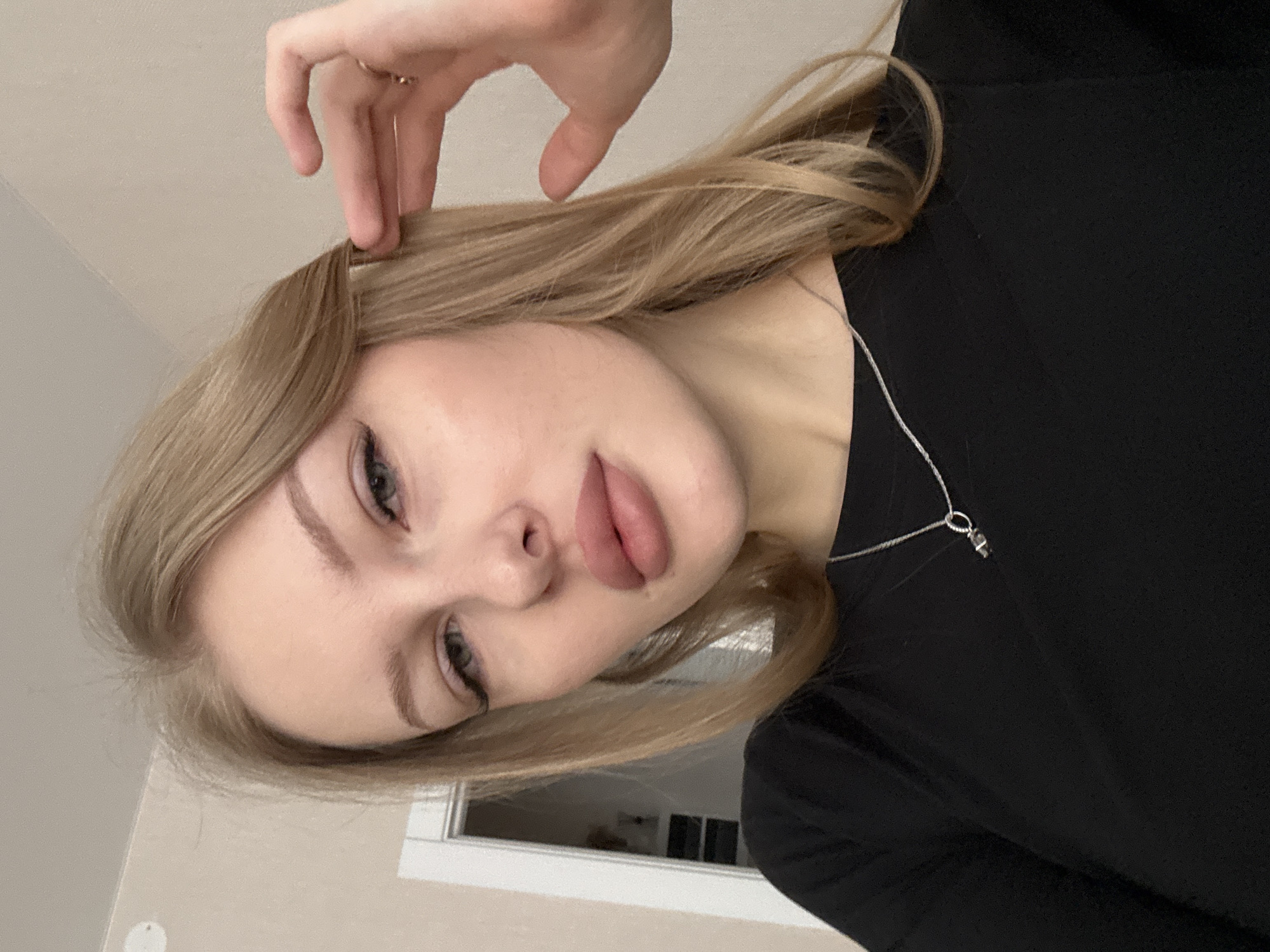 The image displays a close-up indoor portrait of a young adult female, reportedly located in the city of Perm, Russia. The subject, positioned centrally, has light skin, blonde hair, and blue-grey eyes, with prominent eye makeup including winged eyeliner and dark mascara. Her lips appear full, with a matte pink lipstick application. She wears a black, long-sleeved top and a thin silver chain necklace with a dark, possibly cylindrical, pendant. A gold-toned ring is visible on the ring finger of her right hand. Her right hand is raised, with fingers touching the blonde hair near her temple on the right side of the frame. The background consists of a plain light beige wall. To the left, a partially visible white door frame leads into a dimly lit interior, revealing dark shelving or furniture. A portion of a circular white object, possibly a light fixture or smoke detector, is visible on the ceiling in the upper left corner. The subject's gaze is directed towards the camera.