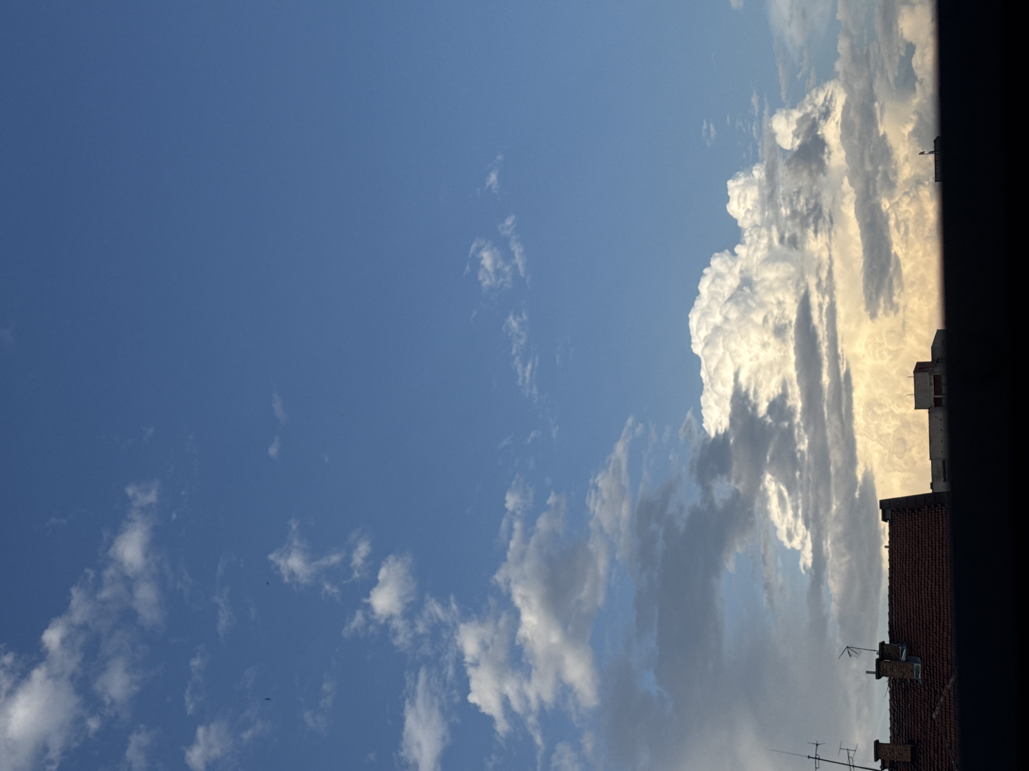 An outdoor, eye-level photograph captures a dynamic sky over a cityscape, likely from a window or balcony in Plovdiv, Bulgaria. The vast expanse of the upper left and central sky is a clear, deep blue with scattered, wispy white clouds. Towards the right side, a substantial formation of bright, voluminous clouds, possibly cumulus or cumulonimbus, dominates the frame. These larger clouds are vividly illuminated with a golden-yellow glow on their edges, suggesting either sunrise or sunset, while other parts appear in shadow, creating strong contrasts.
Along the bottom right and extending up the right edge, parts of urban buildings are visible. The lower right features red-tiled rooftops, complete with chimneys and various antennas, typical of residential or commercial structures. Further to the right, a darker, silhouetted structure, possibly another building or a part of a window frame, casts a stark line against the illuminated clouds. The overall scene suggests a late afternoon or early evening view, characterized by dramatic cloud formations and urban architectural elements. There are no people or discernible text in the image.