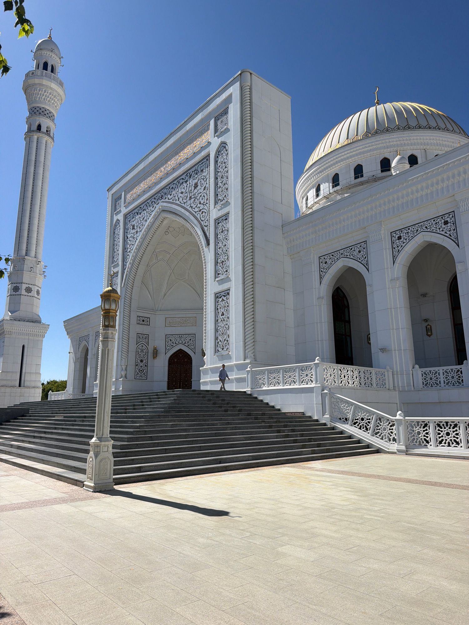 This image depicts the Shali Grand Mosque in Shali, Russia, under a clear blue sky. The mosque is a large, white structure with a prominent golden dome and a tall minaret. The facade features intricate blue and white decorative patterns above arched doorways and windows. A wide, dark-colored staircase leads up to the main entrance, flanked by white ornate railings. In the foreground, a paved plaza is visible, with a lamppost casting a long shadow. A single person, dressed in light-colored clothing, is walking up the stairs on the right side of the mosque. The environment appears to be bright and sunny.