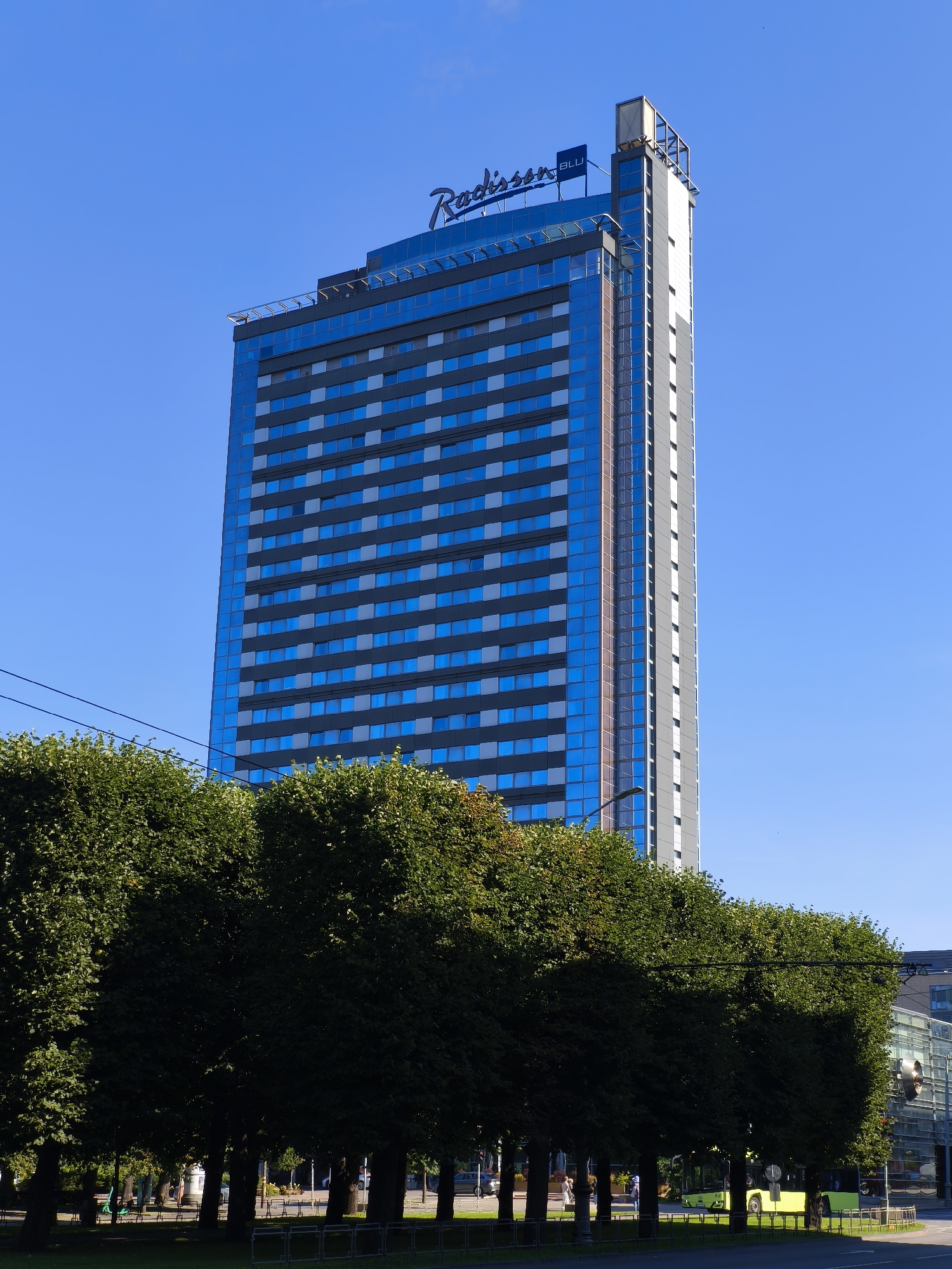 The image shows a modern, glass-facade skyscraper, identified by the "Radisson Blu" logo at its apex, situated in Riga, Latvia. The building has a predominantly blue glass exterior with a grid of lighter blue and grey square windows. To the right of the blue glass section, the building's facade transitions to a grey, more angular design. In the foreground, a dense row of mature, leafy green trees partially obscures the lower portion of the skyscraper. Beyond the trees, a street is visible with a bright yellow bus, a few pedestrians, and streetlights. The sky above is a clear, vibrant blue. The presence of the Radisson Blu hotel suggests an urban setting, likely in a commercial or downtown area of Riga.