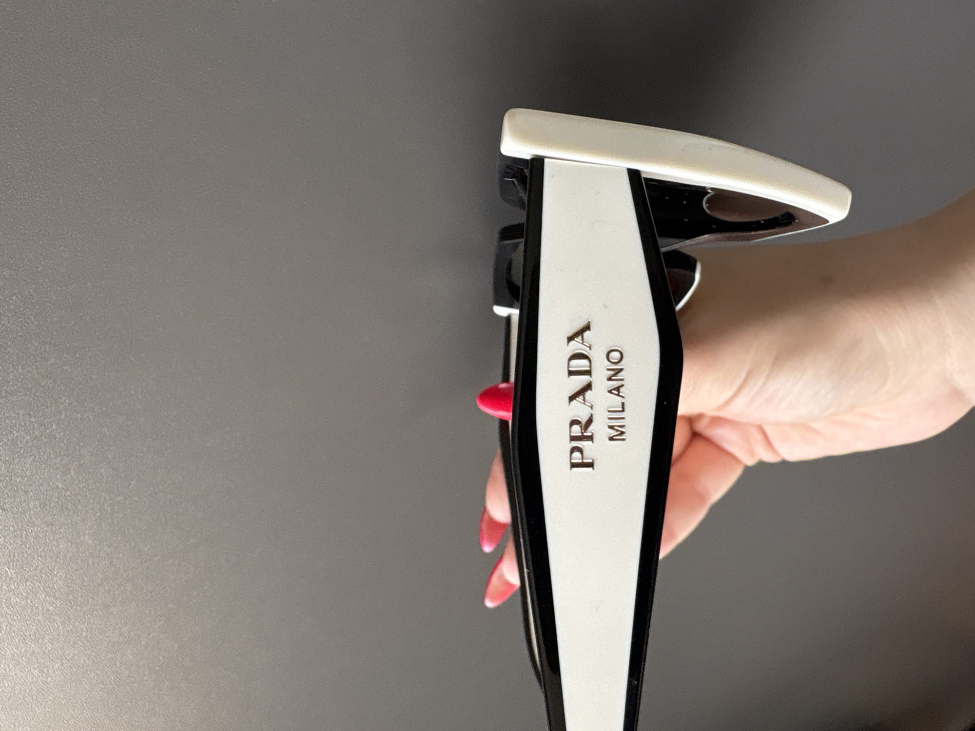 The image shows a pair of Prada Milano sunglasses held in a person's hand. The sunglasses have a white and black frame with the "PRADA MILANO" logo printed vertically on the side. The background is a plain gray surface. The location is not indicated in the image, and is impossible to be in Cheshnegirovo, Bulgaria.