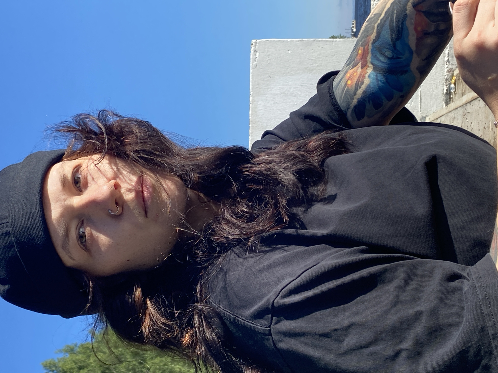 A medium shot captures a young woman with long, dark, wavy hair and brown eyes, wearing a black beanie and a black t-shirt. She has a nose ring and a tattoo on her left arm. The woman is outdoors, with a bright blue sky in the background. To her left, a large white structure with a textured surface is visible. The scene appears to be a waterfront area in Chaikovsky, Russia, with trees on the left and a hint of water and a distant boat on the right. The lighting suggests it is daytime.