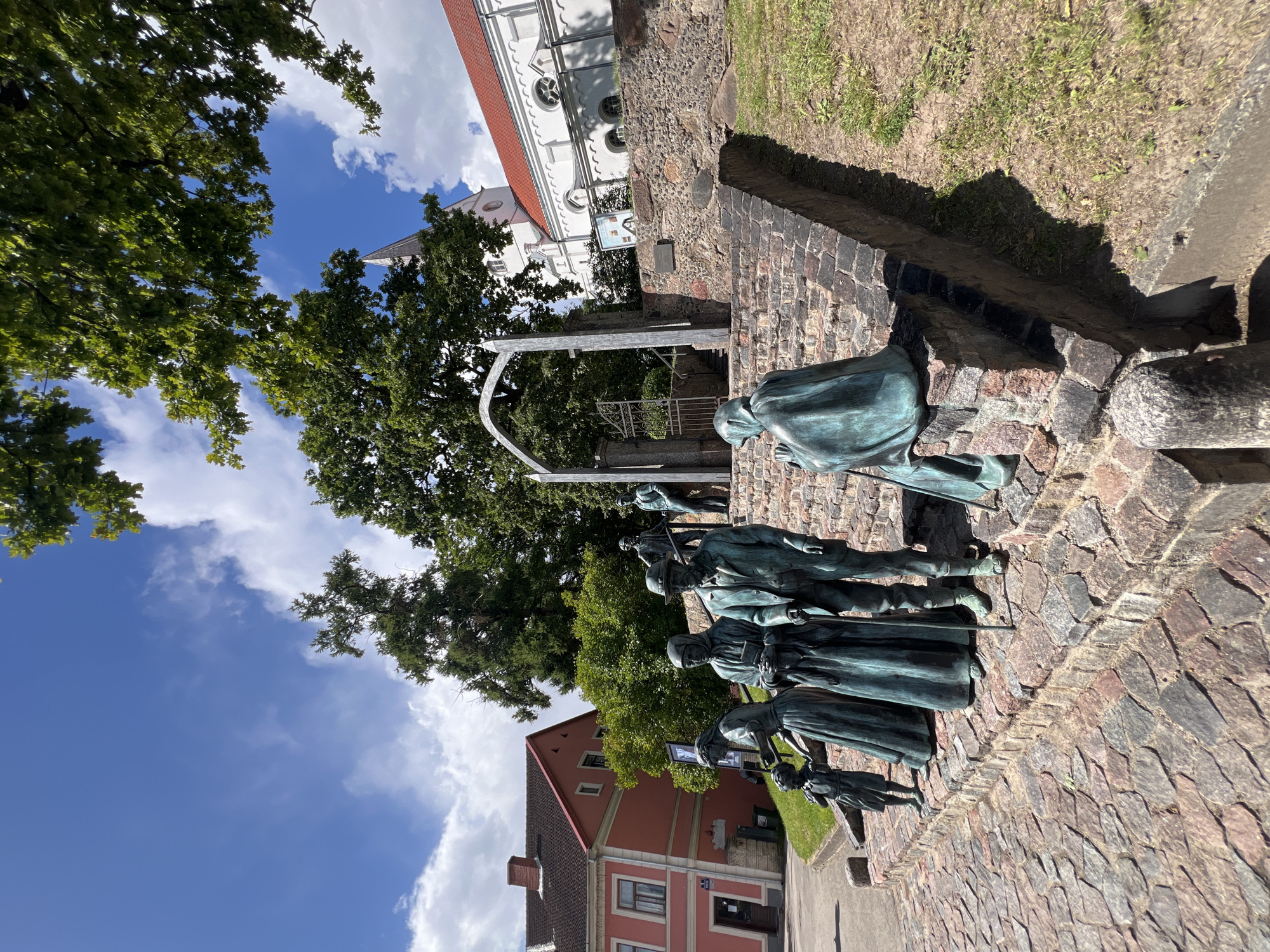 This is a low-angle, outdoor, eye-level shot of a bronze sculpture depicting a family group of several people, possibly representing a historical event or a typical family from the past. The sculptures are located in front of a white church with a red roof, which is situated behind a stone wall and a small grassy embankment. In the background, there is a pink building and a cobblestone street. The sky is blue with white clouds, and there are trees with green foliage in the foreground and background. The sculptures include a child, several adults dressed in traditional clothing, and at least one person appearing to be praying. The overall scene suggests a public space, likely in a town or city square, possibly a memorial or a tourist attraction in Saldus, Latvia.