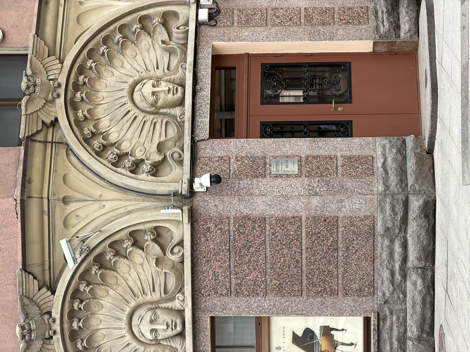 The image depicts the facade of a building in Saint Petersburg, Russia, specifically showcasing architectural details. Two circular bas-relief sculptures, each featuring a human face surrounded by radiating patterns, are positioned vertically on the left. An owl sculpture is on either side of the circles. Adjacent to these sculptures is a section of the building's exterior, composed of brick patterns. A security camera is attached to the building. A brown wooden door with wrought-iron details is present above a placard with Cyrillic text. Below the door and placard, a poster featuring a piano is visible. The background includes a stone wall and an additional window. The overall architectural style suggests a historical or classical influence. The image is taken from a slightly low angle, and the photograph appears to be rotated slightly clockwise.