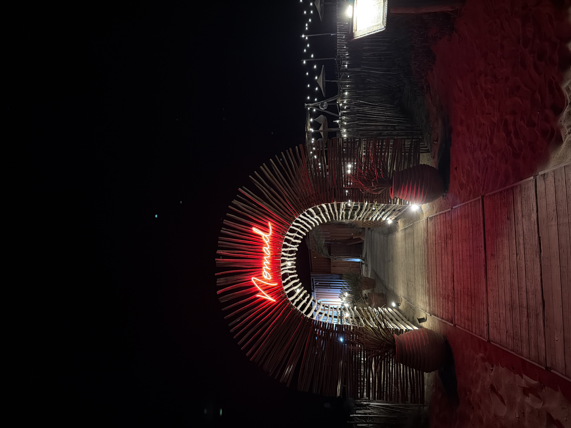 This image depicts the entrance to a establishment called "Nomad" in Torreilles, France, at night. A pathway made of weathered wooden planks leads through a semicircular archway constructed from numerous thin wooden poles. The archway is adorned with a bright red neon sign spelling out "Nomad" in cursive. On either side of the pathway, large, terracotta-colored urns hold palm-like plants. The ground on the sides of the pathway is sand, illuminated by red ambient lighting. Beyond the archway, more wooden fencing and a line of string lights are visible, suggesting an outdoor seating area or beachside establishment. The sky is dark and clear, with a few faint lights visible in the distance. The overall scene conveys a festive and inviting atmosphere, likely a restaurant, bar, or beach club.