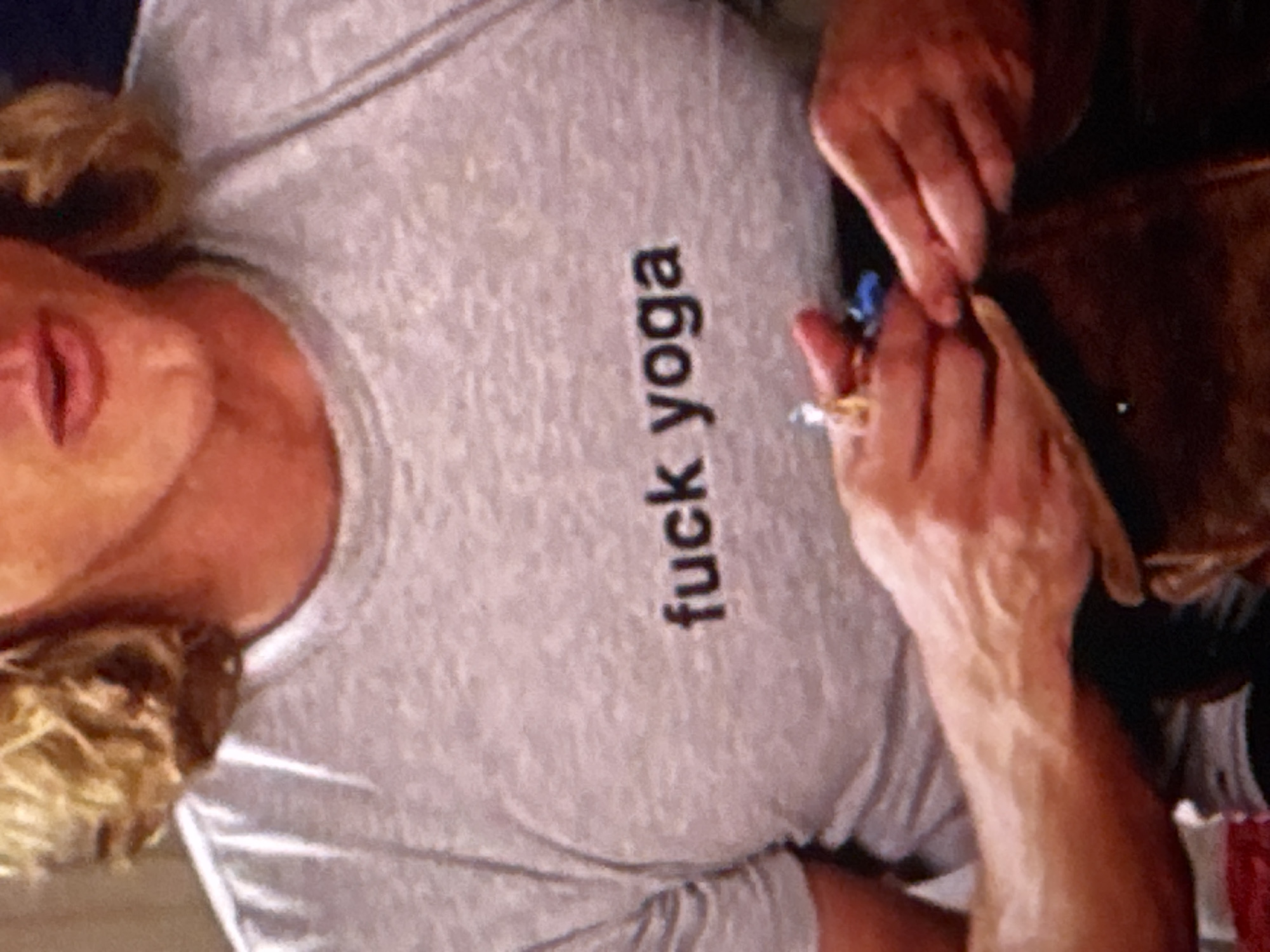 The image is a close-up shot of a person wearing a grey t-shirt with the words "fuck yoga" printed on it in black lettering. The person's chest and hands are visible. They are wearing rings on their fingers. The background is out of focus and appears to be indoors. There is no discernible information about the location in Kuta Selatan, Indonesia, in this image.